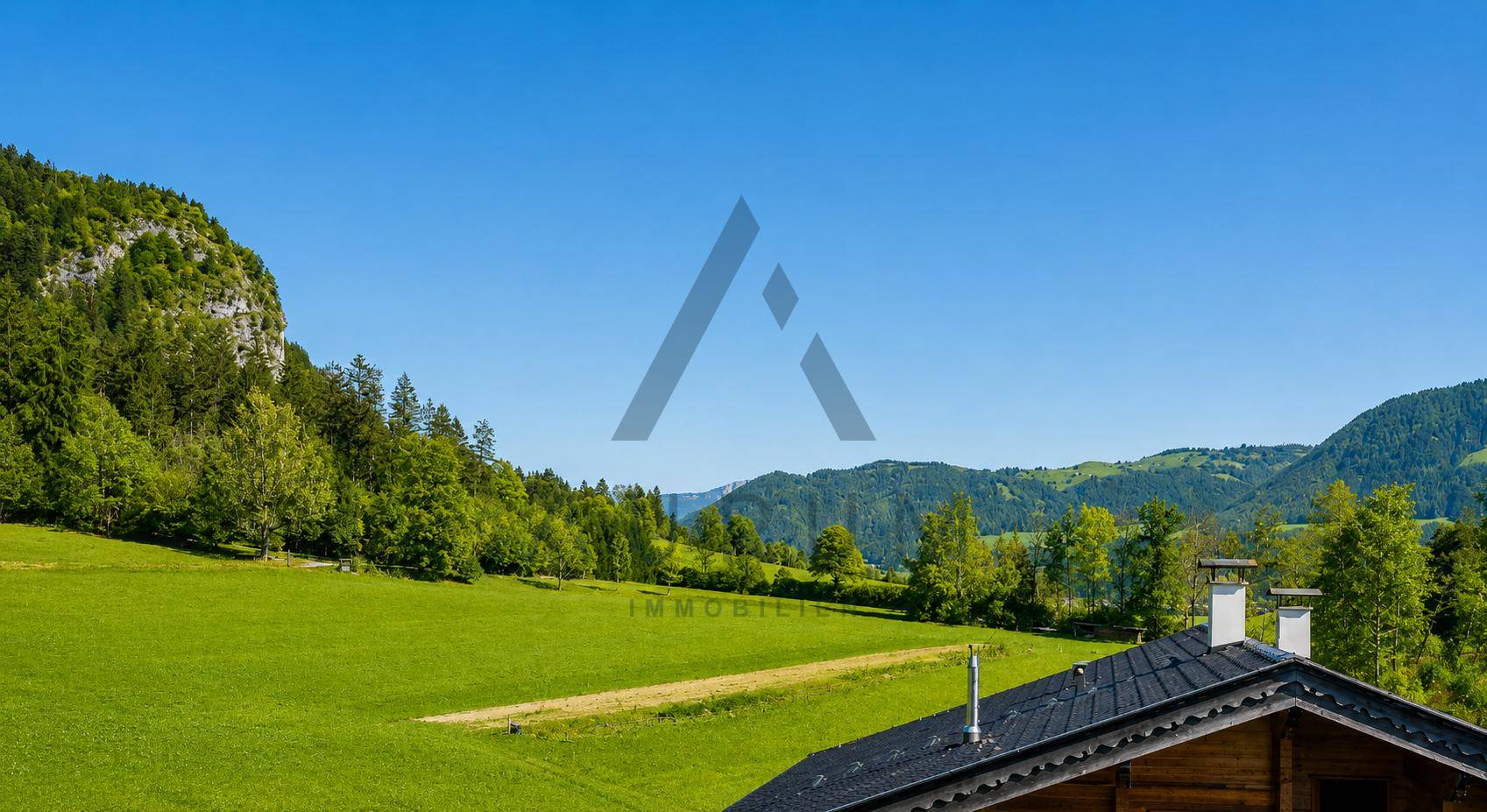 Weitläufige grüne Wiesen und bewaldete Berge unter blauem Himmel, mit einem Hausdach im Vordergrund.