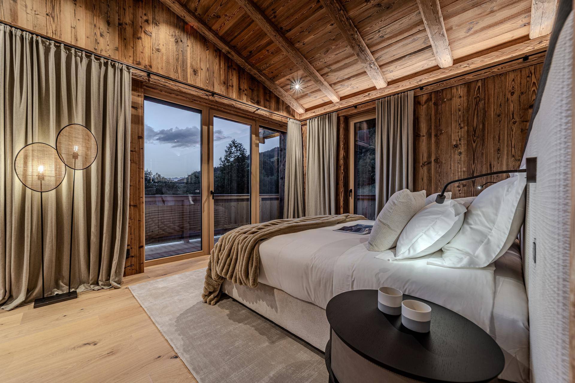 Helles Schlafzimmer mit Holzbalkendecke, Balkonzugang und Abendstimmung mit Bergblick.