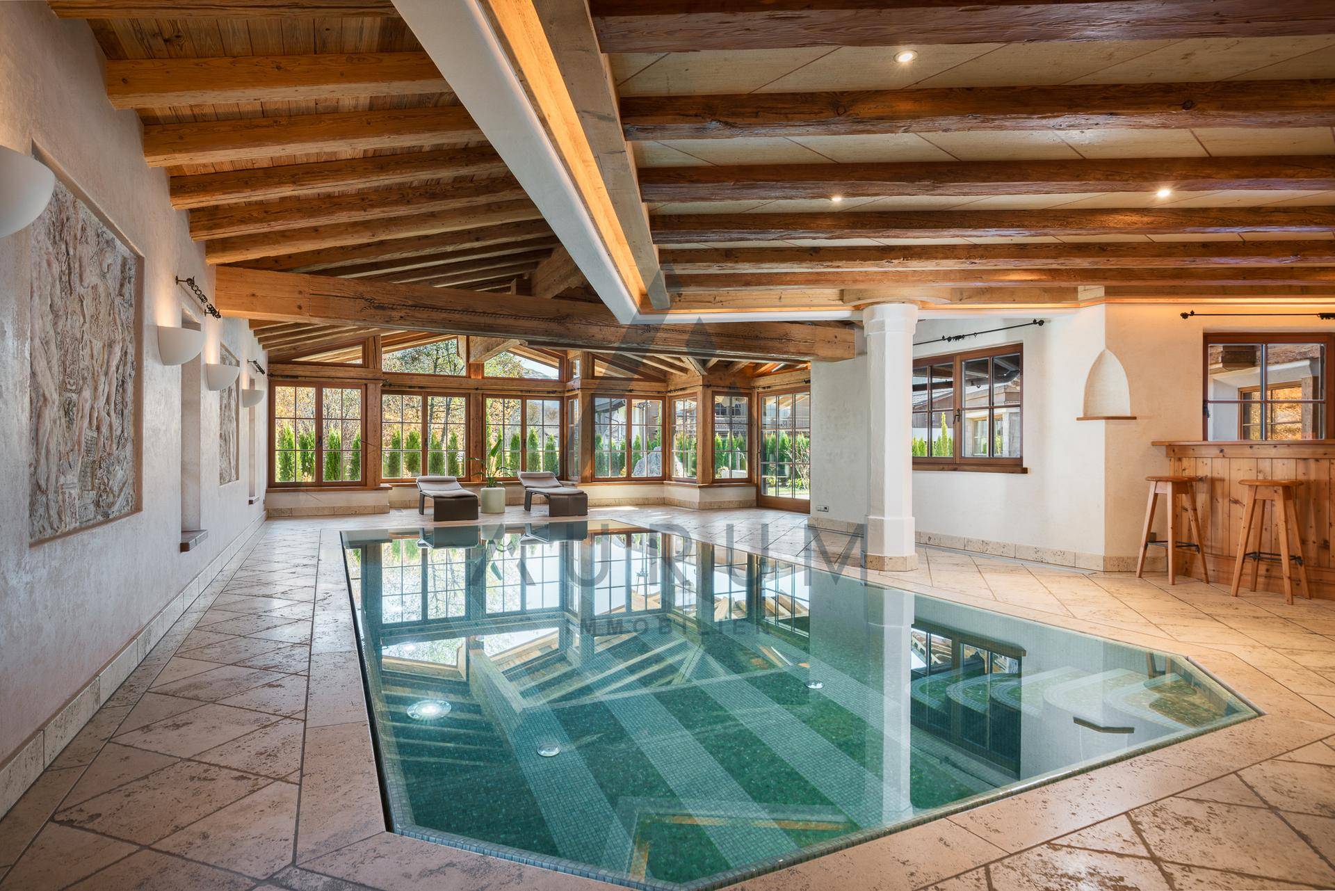 Innenpoolbereich mit großen Fenstern, Holzbalkendecke und Liegemöglichkeiten.