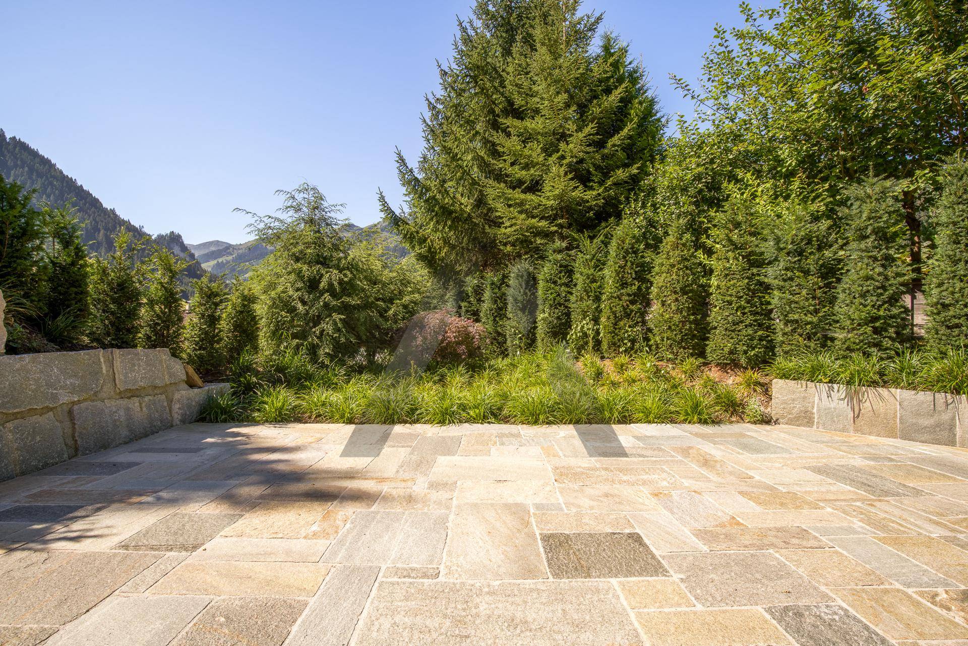 Geräumige gepflasterte Terrasse mit Steinmauern und üppiger Bepflanzung, umgeben von Bergen.