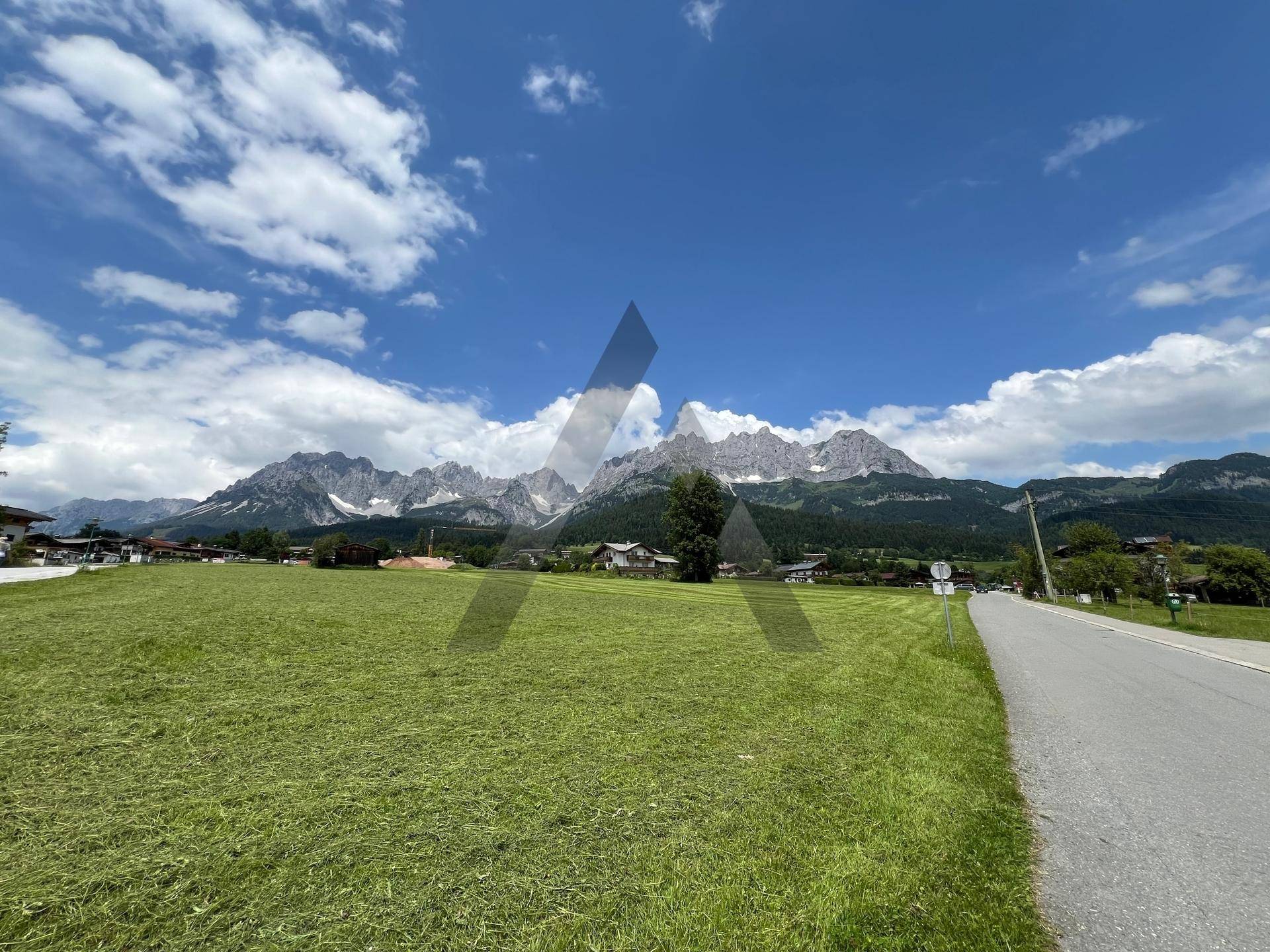 Weitläufige Landschaft mit einer Straße, grünen Feldern und einer beeindruckenden Bergkulisse unter blauem Himmel.