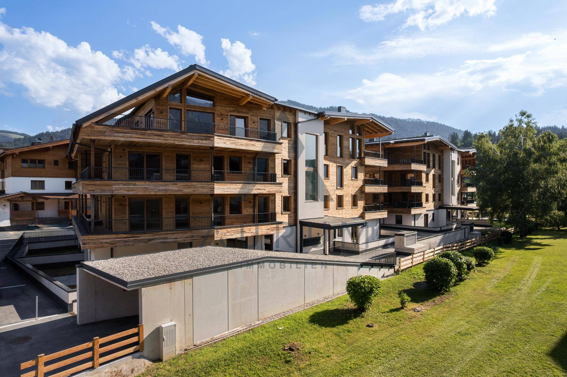 Modernes Holzgebäude mit Tiefgaragenzufahrt und weitläufiger Grünfläche in alpiner Umgebung.