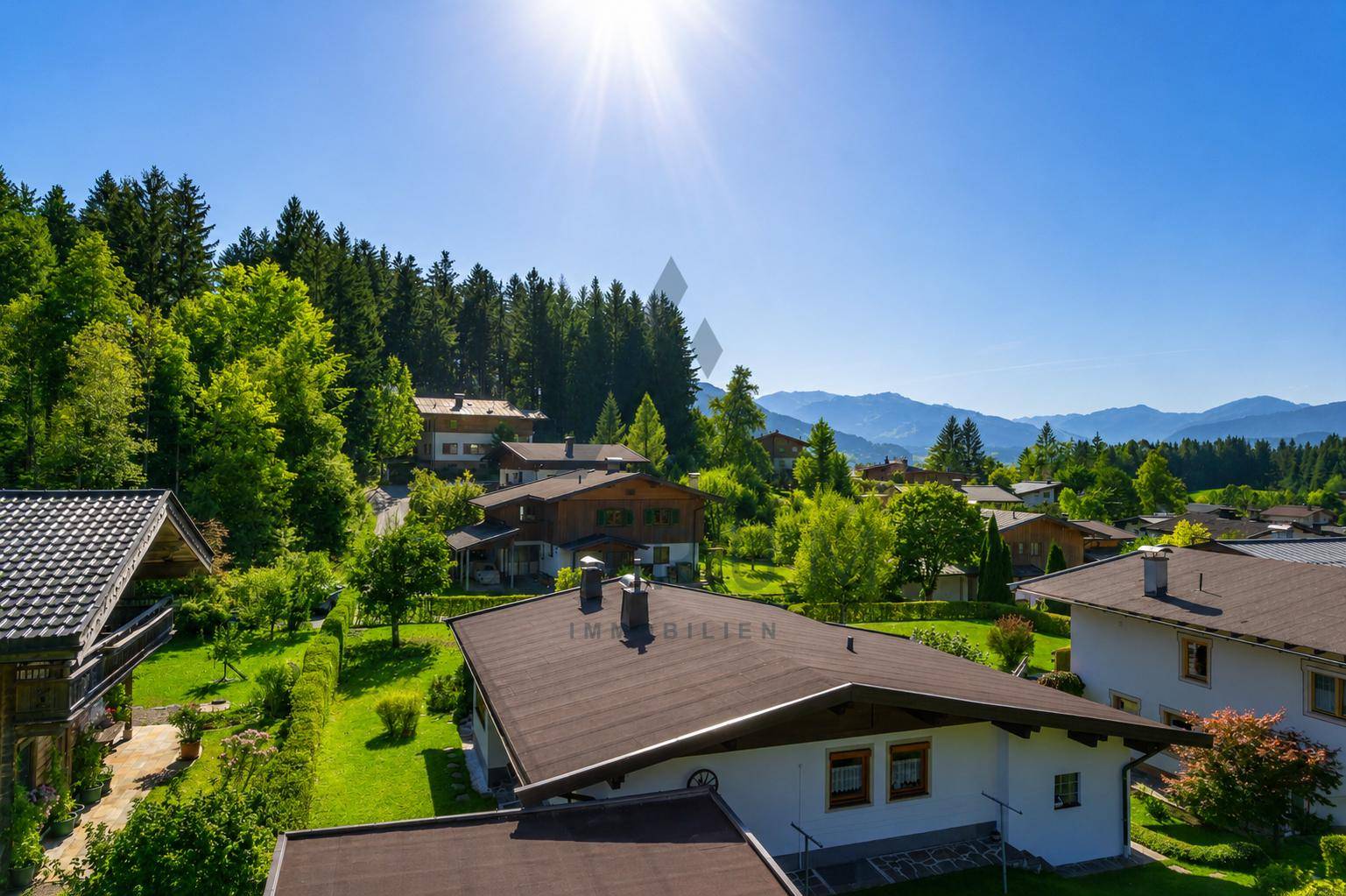 Sonnige Luftaufnahme einer Wohnsiedlung umgeben von Wäldern und Bergen in der Ferne.