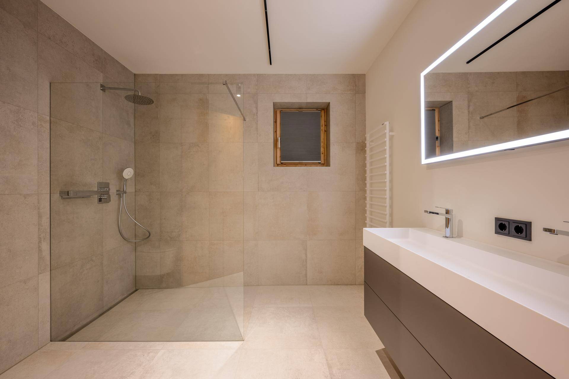 Modernes Badezimmer mit begehbarer Dusche, großformatigen Fliesen und beleuchtetem Spiegel.