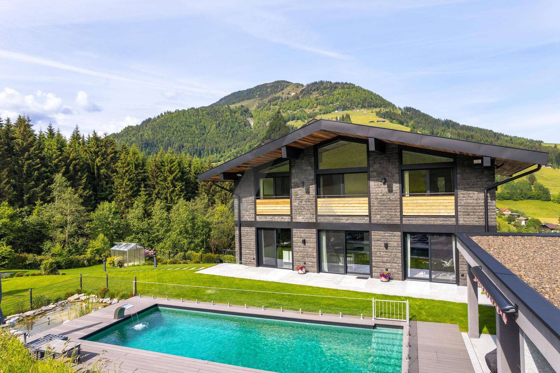 Modernes Haus mit großem Pool und Holzdeck, umgeben von Wald und Bergen.