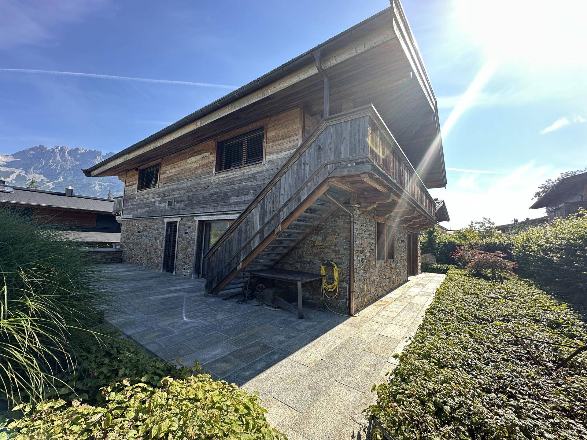 Rückseite des Hauses mit großer gepflasterter Terrasse und Zugang zum Garten, umgeben von üppigem Grün.