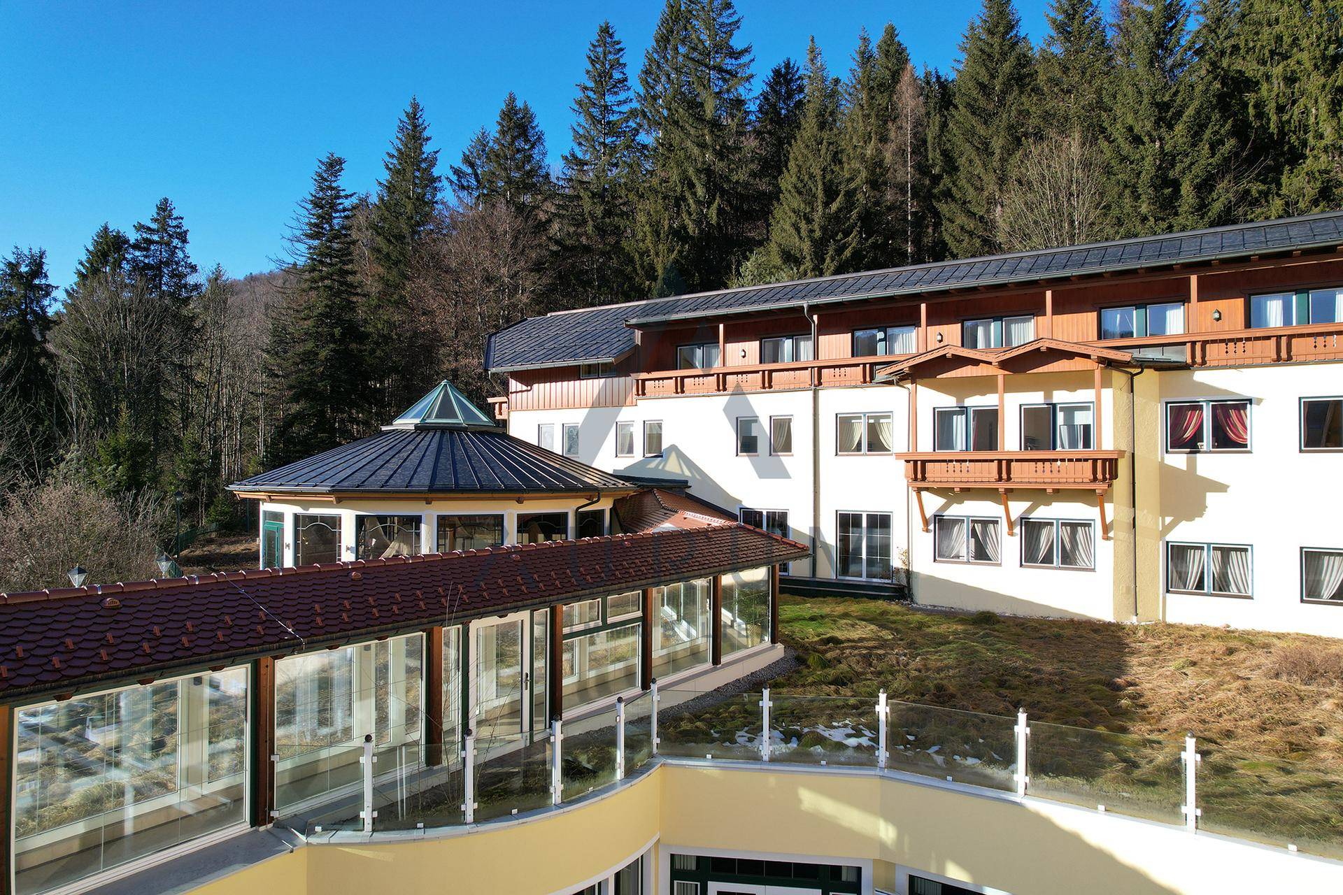 Gebäudefassade mit einem verglasten Gang, einem runden Pavillon mit Kegeldach und Balkonen vor bewaldetem Hintergrund.