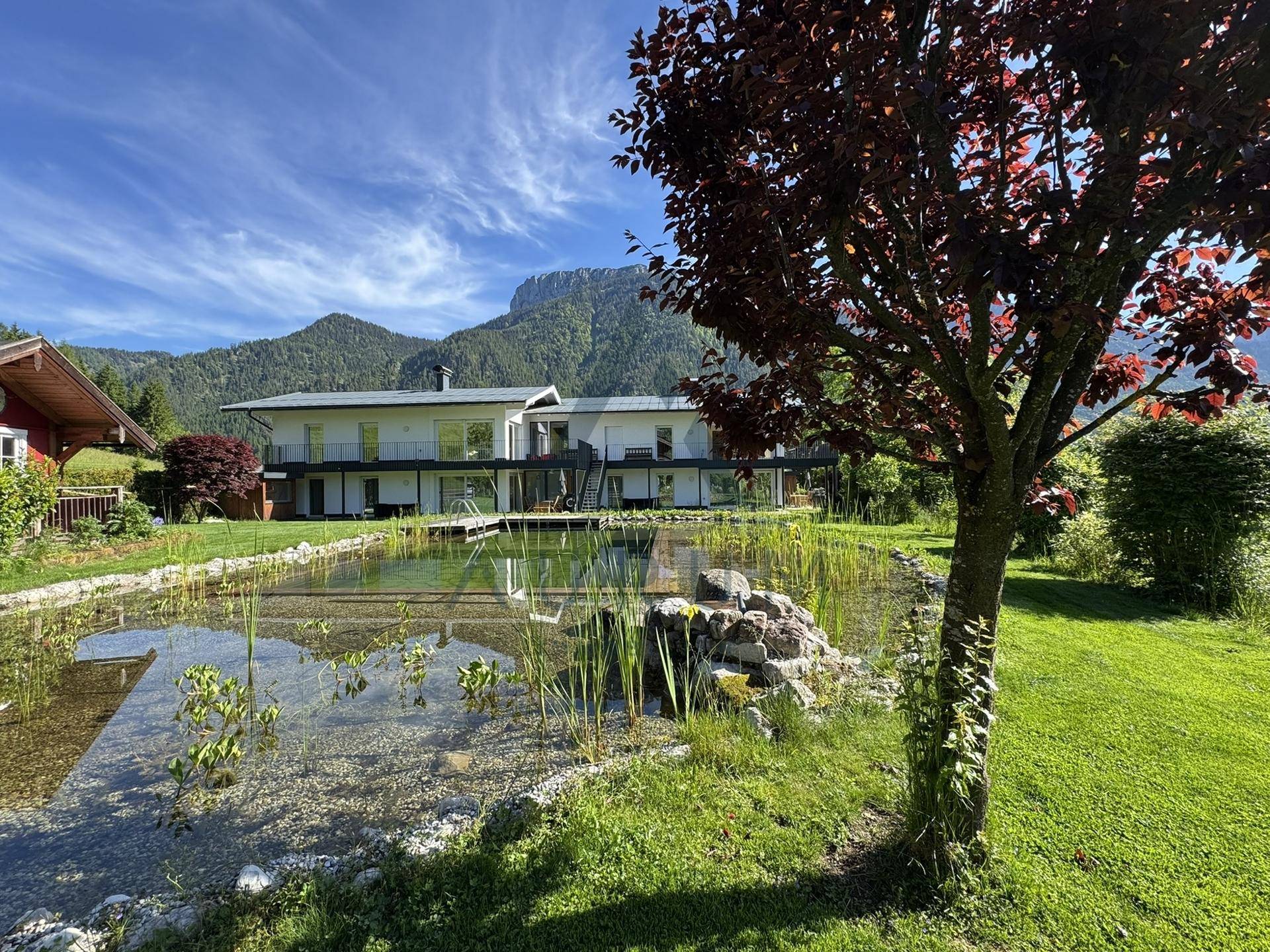 Gepflegter Garten mit Naturteich und Blick auf das moderne Gebäude vor einer Bergkulisse.