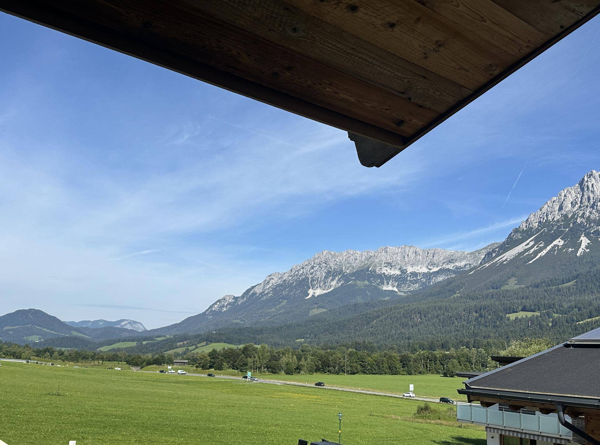 Weitläufiger Blick über grüne Felder und eine Landstraße mit majestätischen Bergen im Hintergrund.