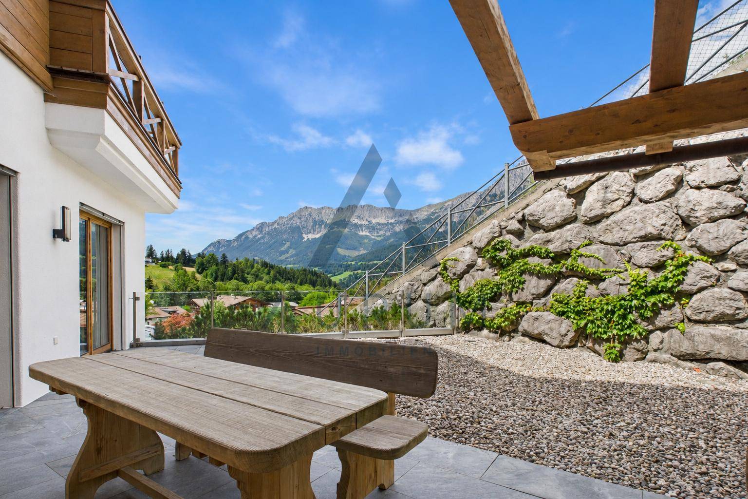 Rustikale Terrasse mit Holztisch und Bänken, umgeben von einer Steinmauer und Bergblick.