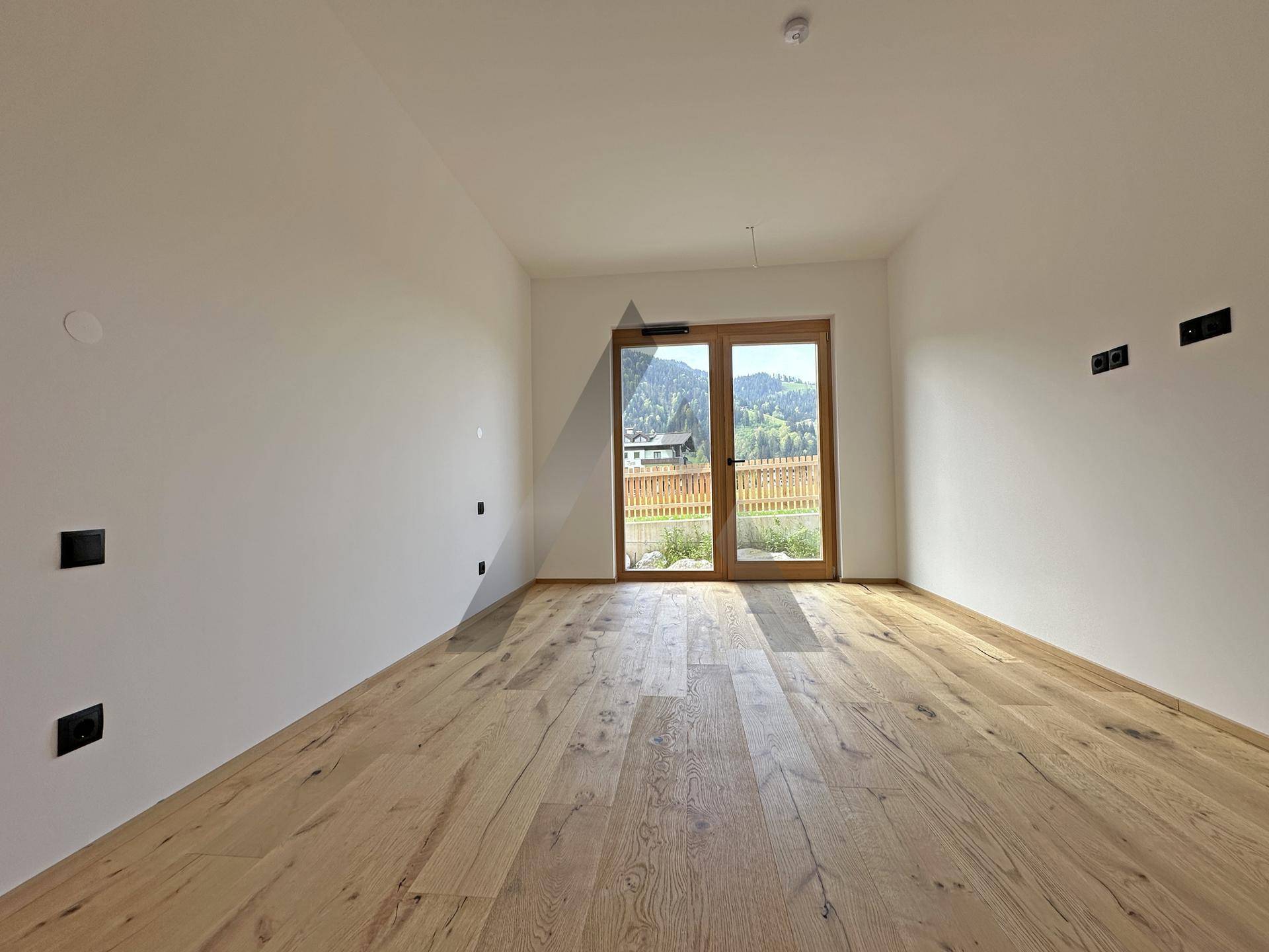 Freundliches Zimmer mit Holzboden und Fenstertür, die einen Blick auf die grüne Umgebung freigibt.