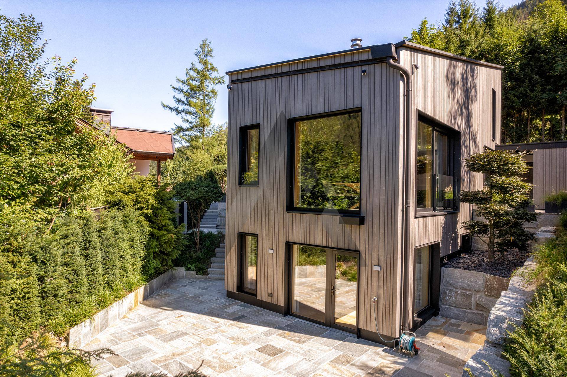 Moderne Holzfassade des Hauses mit großen Fenstern und einer gepflasterten Terrasse im Grünen.
