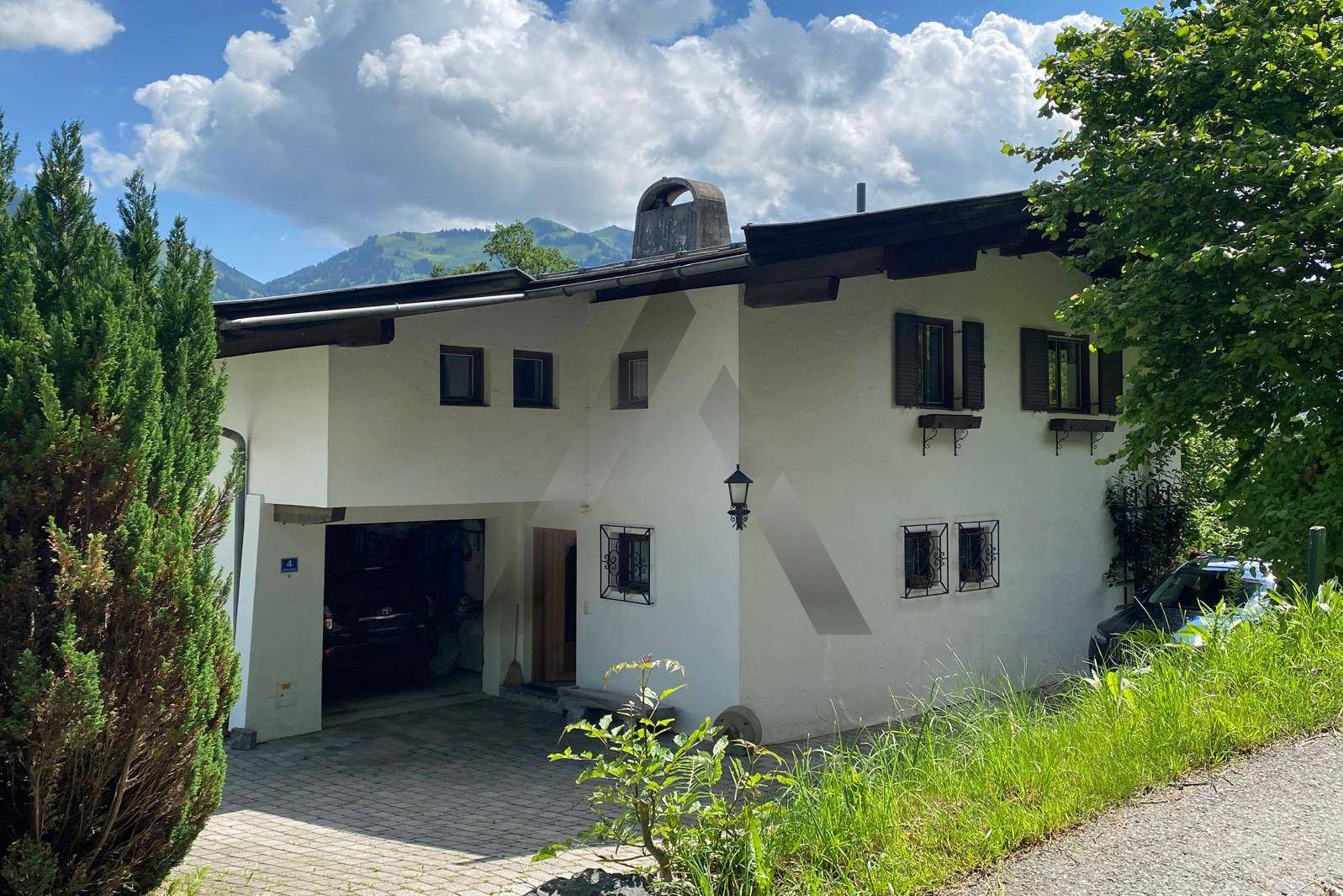 Traditionelles Haus mit weißer Fassade, kleinen Fenstern und integrierter Garage an einem Hang.