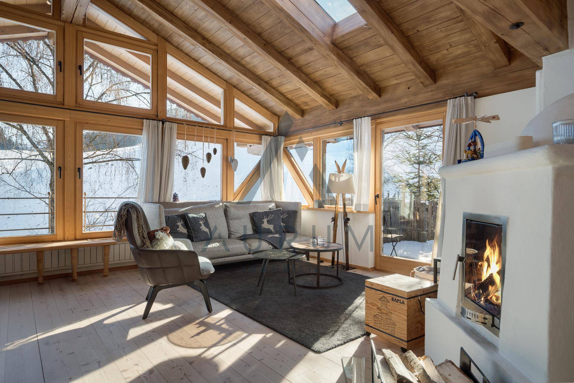 Gemütliches Wohnzimmer mit Kamin, Holzboden und großen Fenstern, die viel Tageslicht hereinlassen.