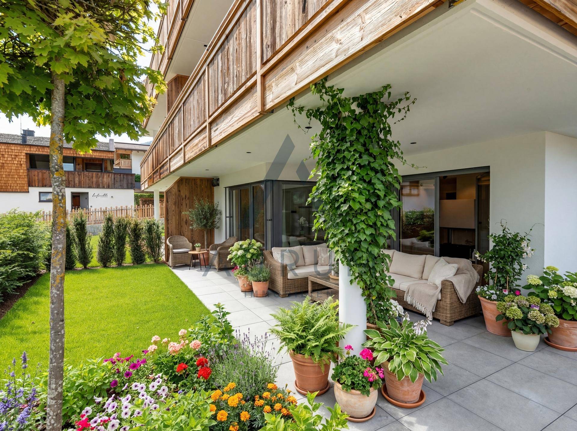 Geräumige Terrasse mit Gartenmöbeln, Rasenfläche und üppigen Blumenbeeten vor einem modernen Gebäude.