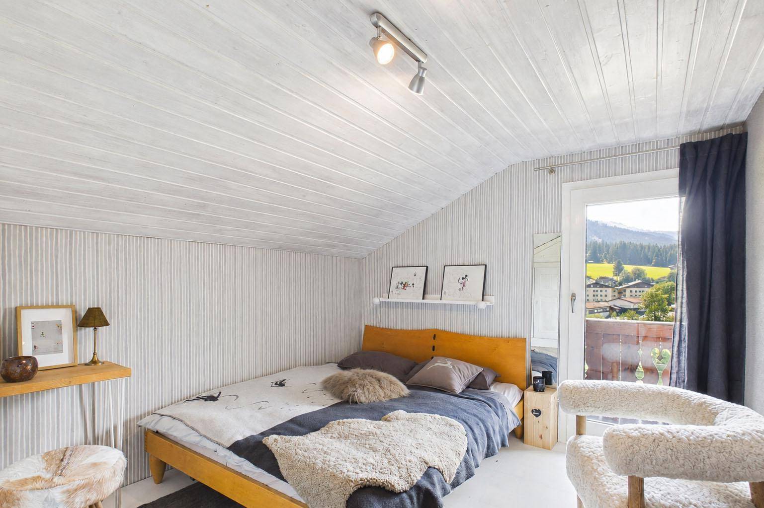 Schlafzimmer im Dachgeschoss mit Holzbett, weißer Holzdecke und Balkonzugang mit Bergblick.