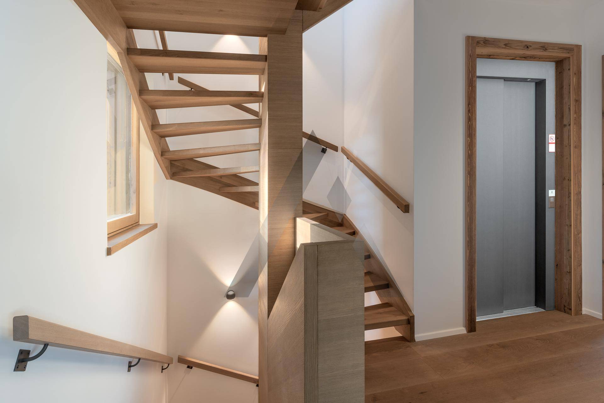 Heller Treppenaufgang mit Holzstufen und Glasgeländer, daneben ein moderner Aufzug.