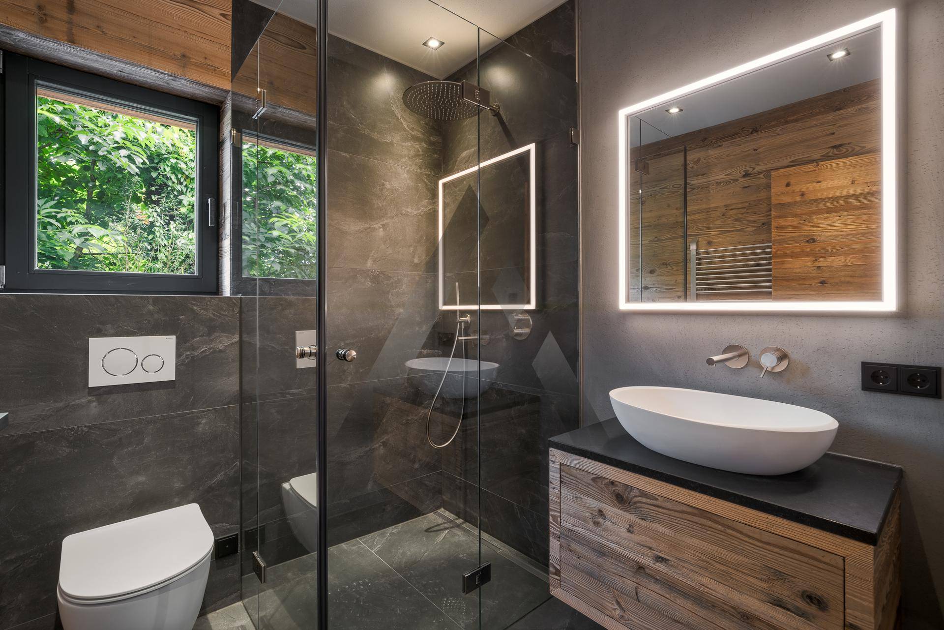 Modernes Badezimmer mit dunklen Fliesen, Holzdetails, Dusche und beleuchtetem Spiegel, mit Fenster.