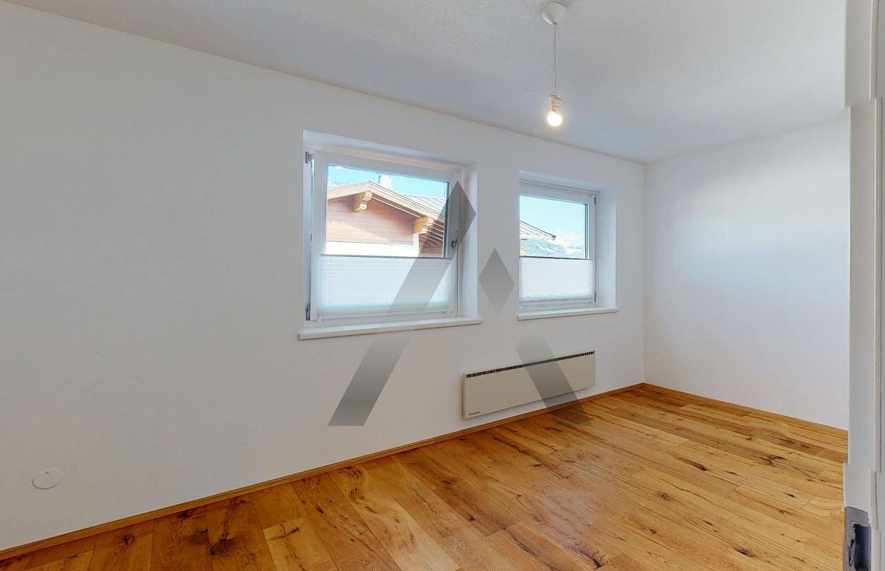 Leeres Schlafzimmer mit Holzboden und zwei Fenstern, die viel Tageslicht hereinlassen.