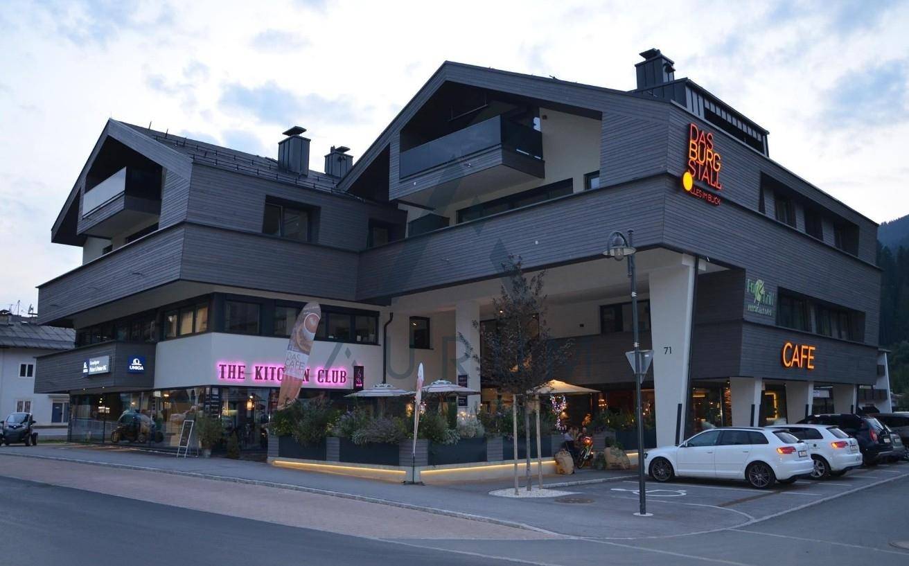 Modernes Geschäftsgebäude am Abend mit verschiedenen Restaurants und Cafés im Erdgeschoss und geparkten Autos davor.