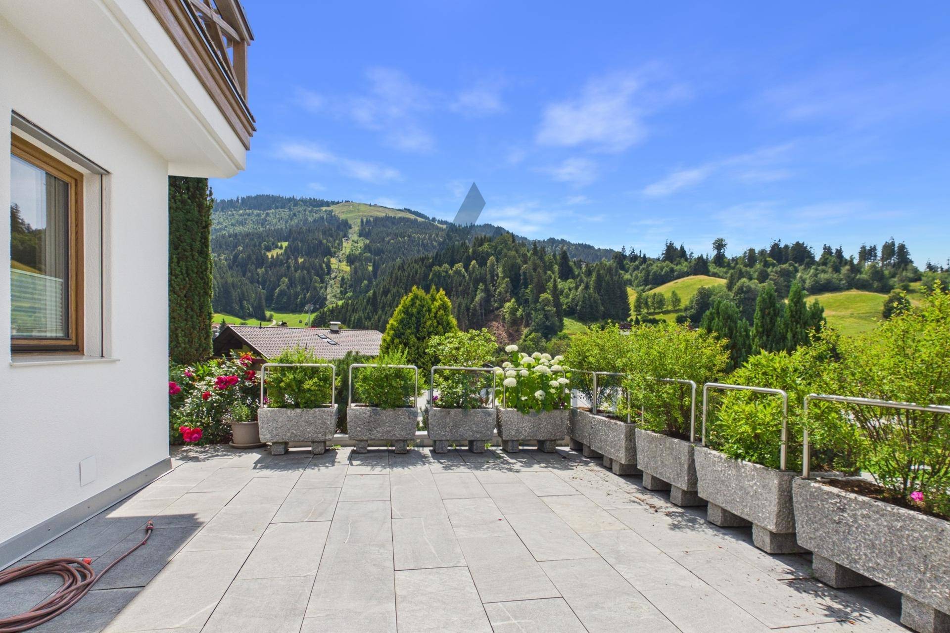 Sonniger Terrassenbereich mit Steinplatten, Blumen und Blick auf die grüne Hügellandschaft.