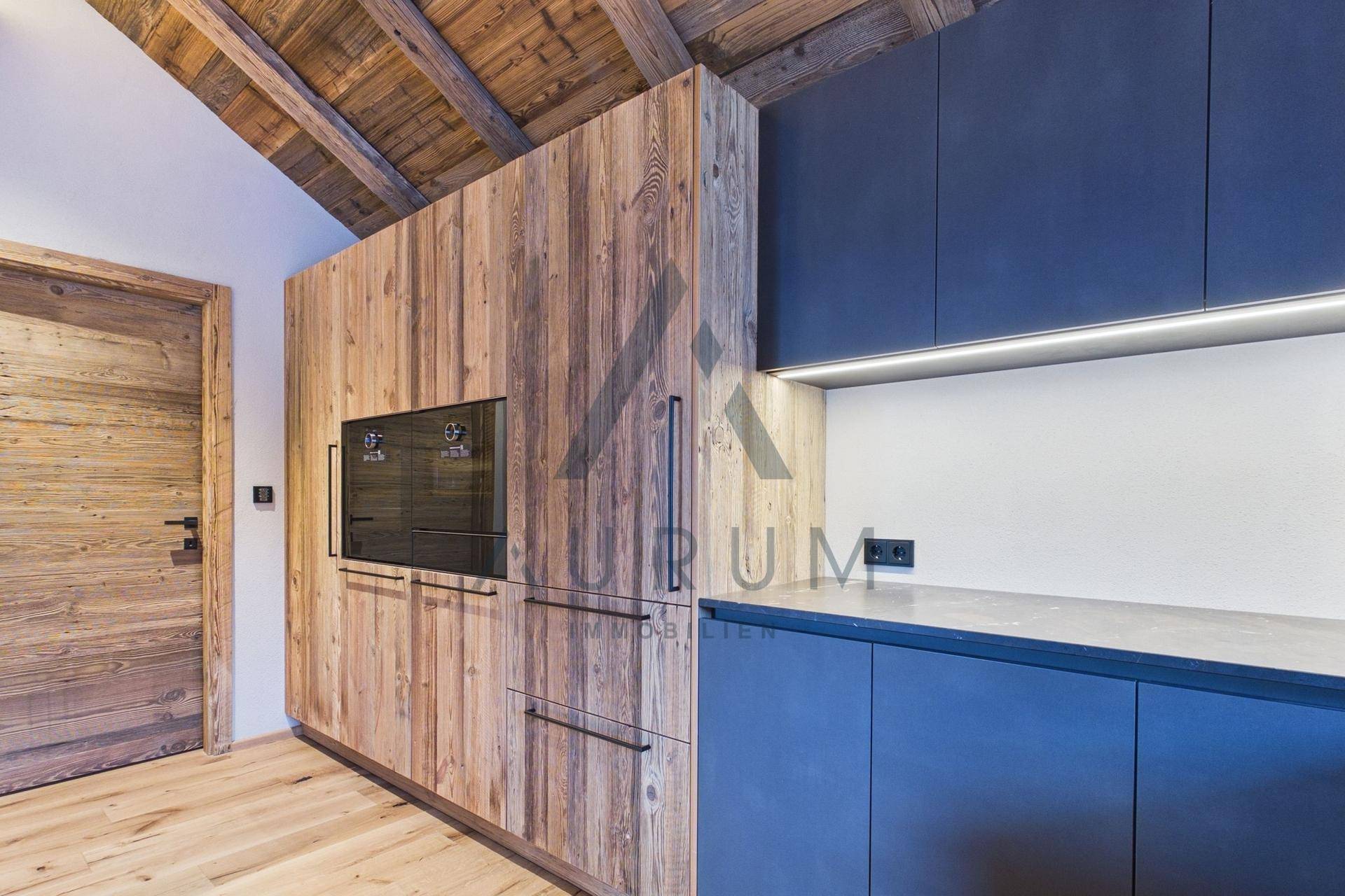 Integrierte Küche mit Holz- und dunklen Fronten, Backofen, Kühlschrank und Holzbalkendecke.