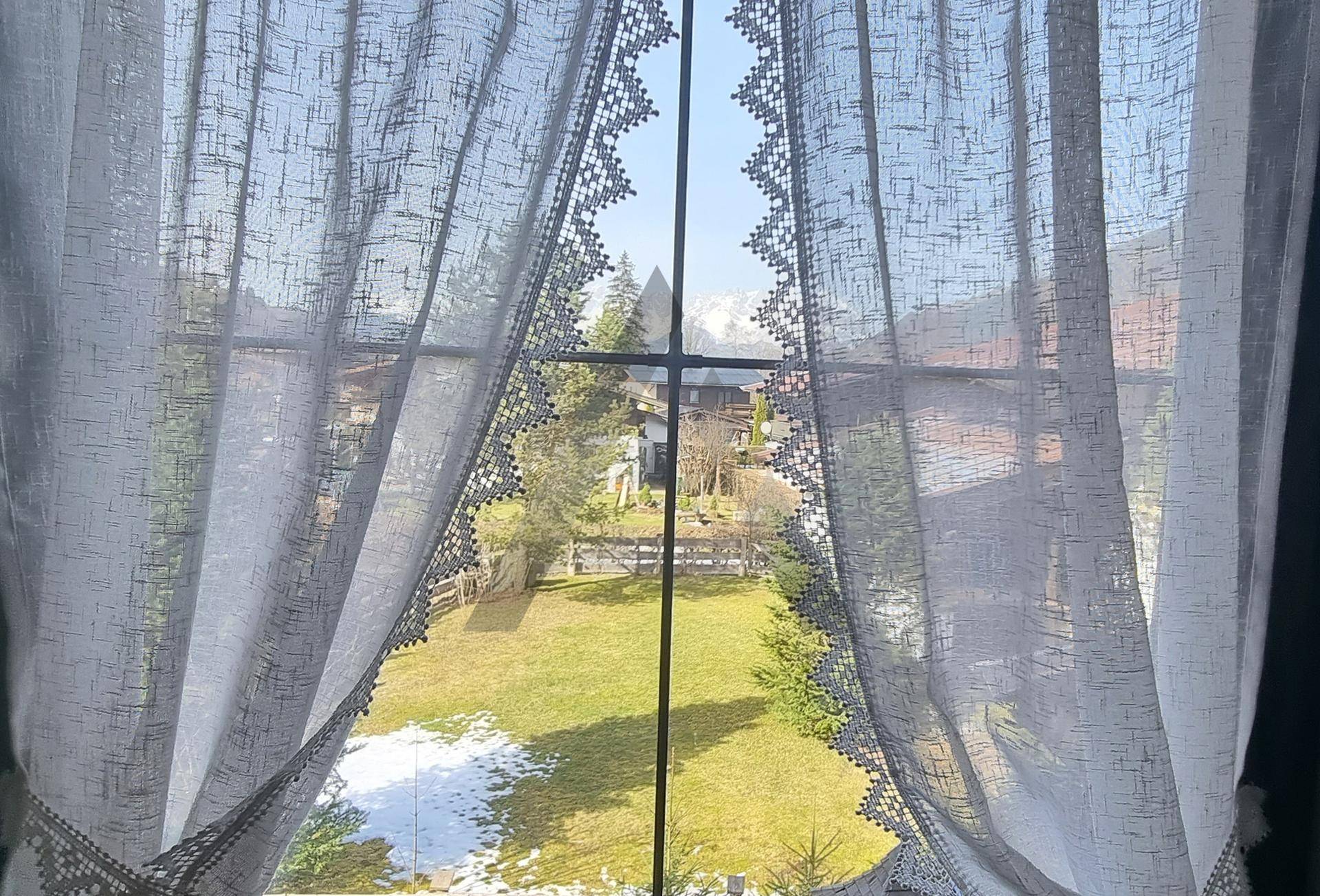 Blick durch ein Fenster mit Spitzenvorhängen auf einen Garten mit Restschnee und die ländliche Umgebung.
