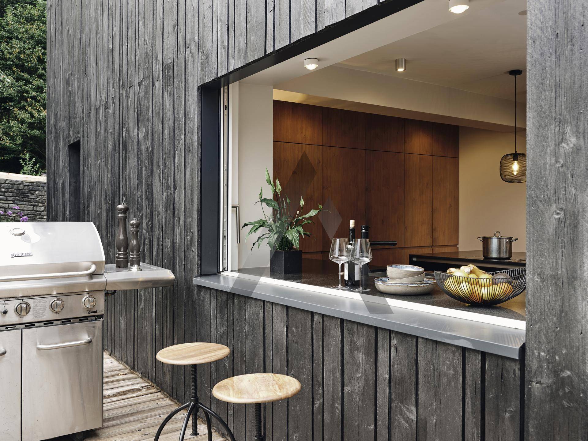 Außenküche mit Grill und Barhocker, verbunden mit dem Innenbereich durch eine große Fensteröffnung.