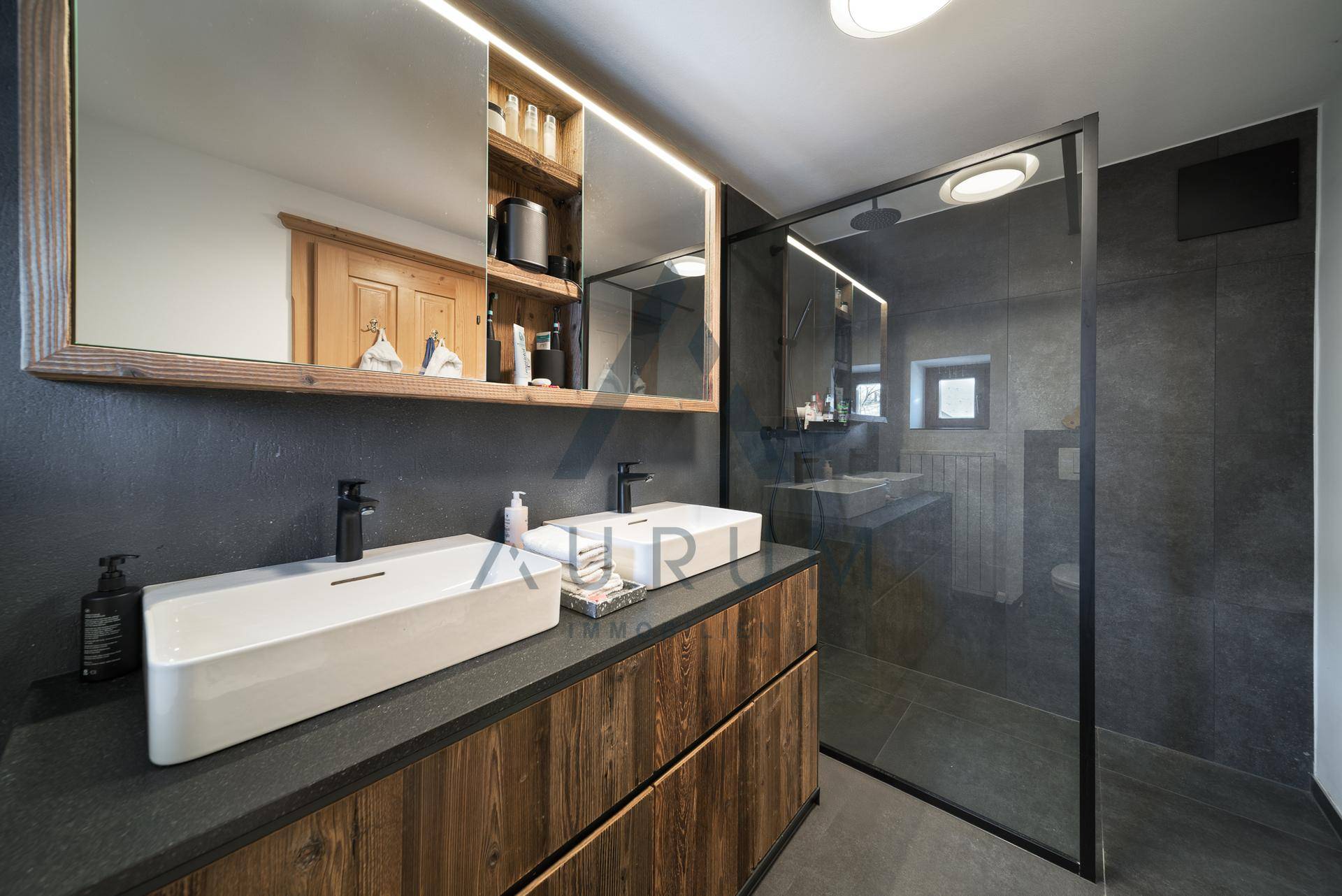 Modernes Badezimmer mit Doppelwaschbecken, großer begehbarer Dusche und dunklen Fliesen.