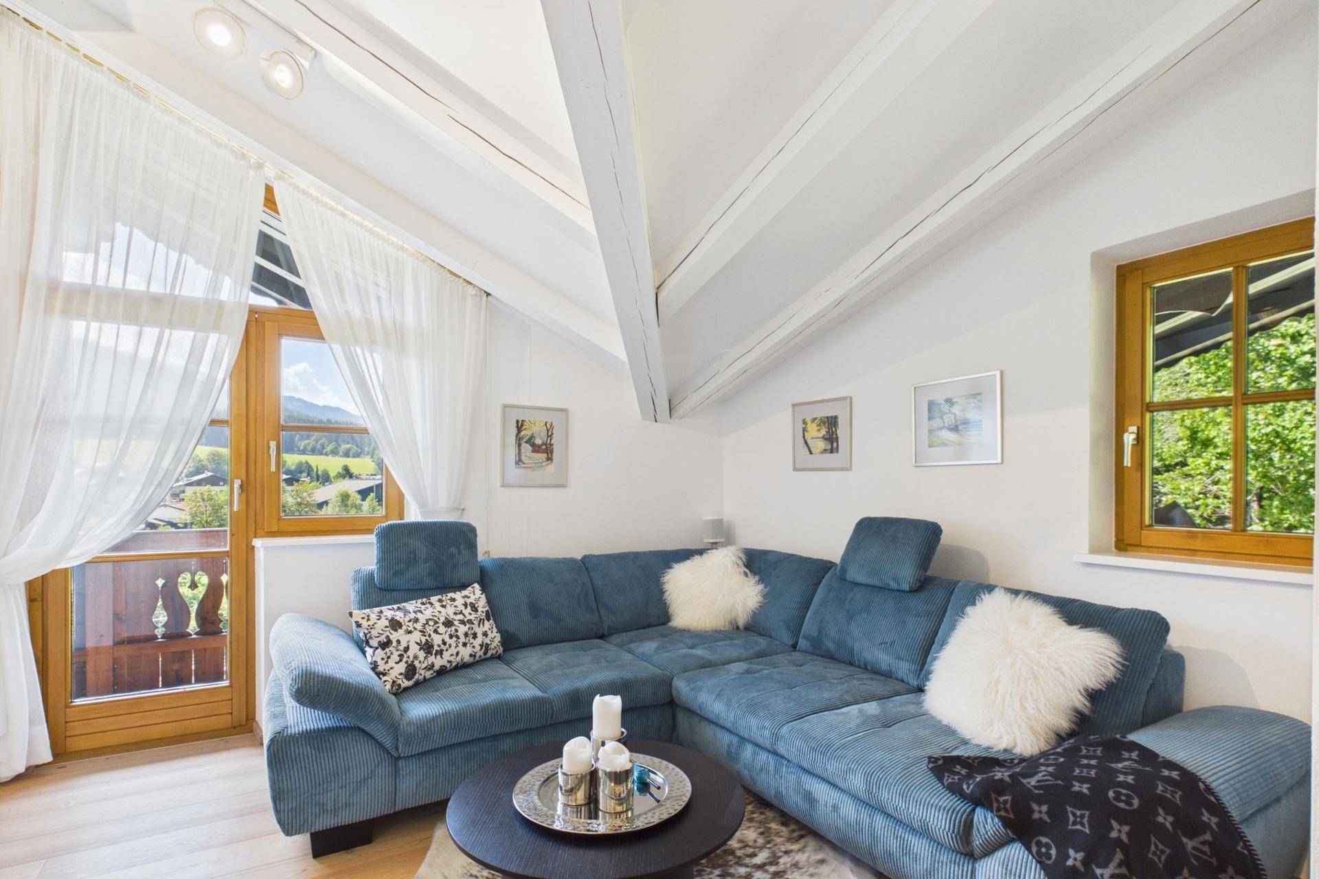 Helles Wohnzimmer im Dachgeschoss mit blauer Couch, Holzbalken und Zugang zum Balkon mit Bergblick.