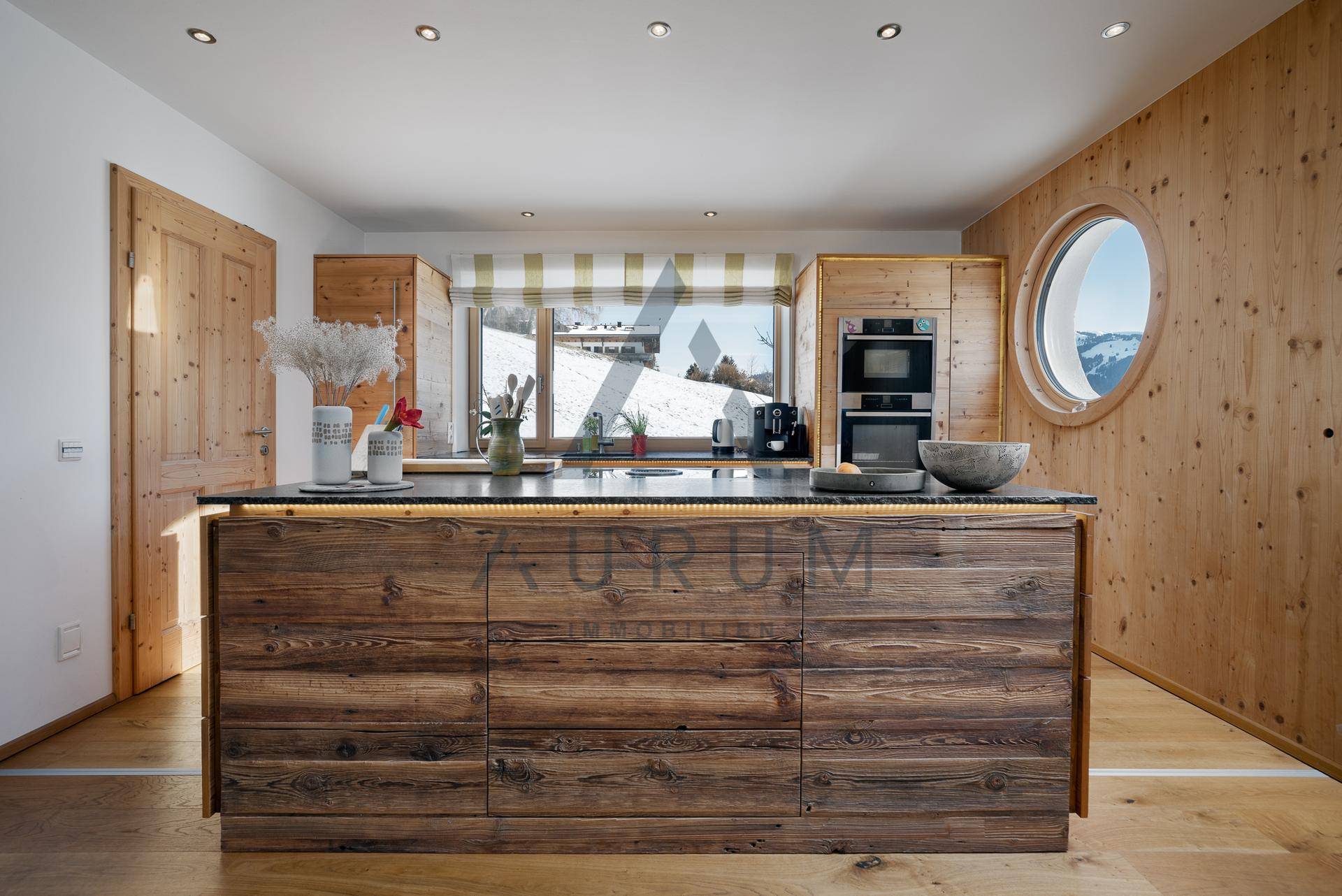 Moderne Holzküche mit Kücheninsel, integrierten Geräten und einzigartigem runden Fenster mit Bergblick.