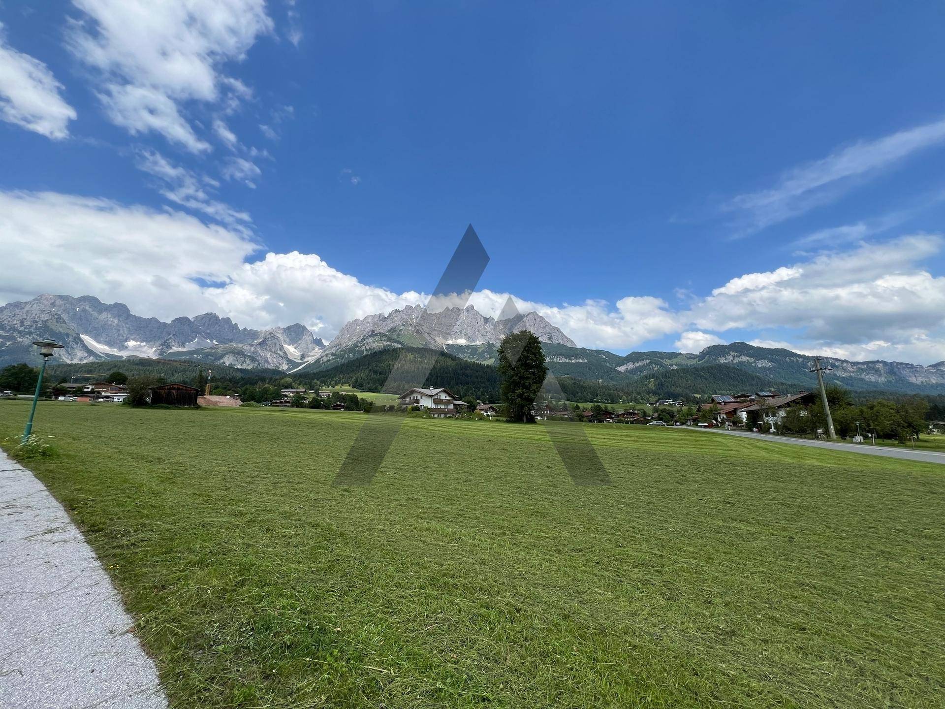 Grüne Wiesen mit einem Spazierweg im Vordergrund und einer malerischen Berglandschaft in der Ferne.