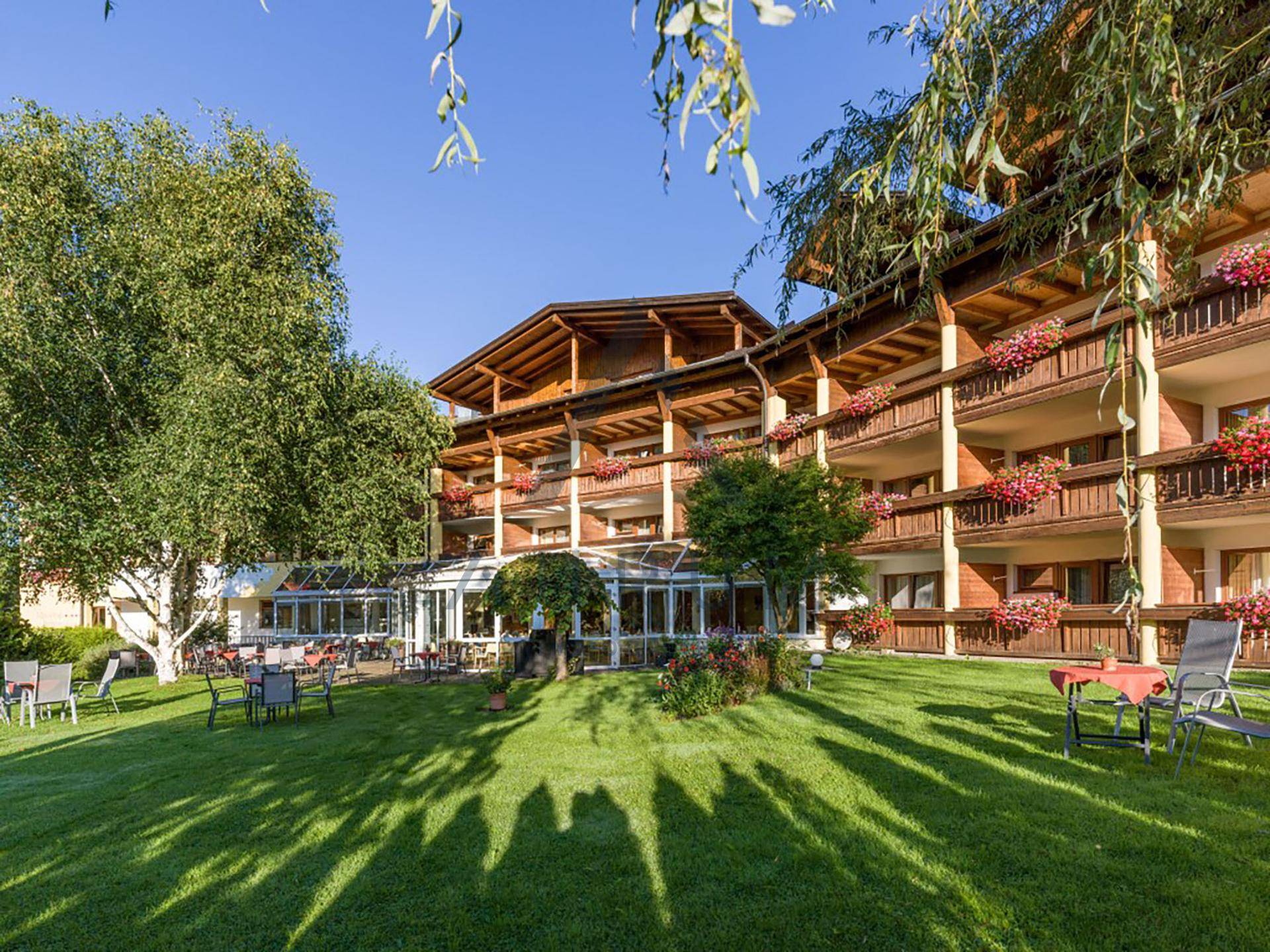 Gepflegte Gartenanlage mit schattenspendenden Bäumen und einem traditionellen Hotelgebäude im Hintergrund.