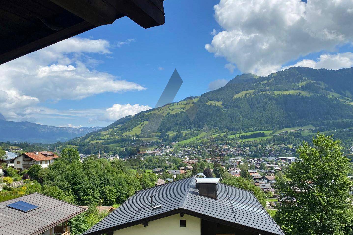 Panoramablick von einem Balkon über Dächer, ein Tal und die umliegende Berglandschaft unter blauem Himmel.