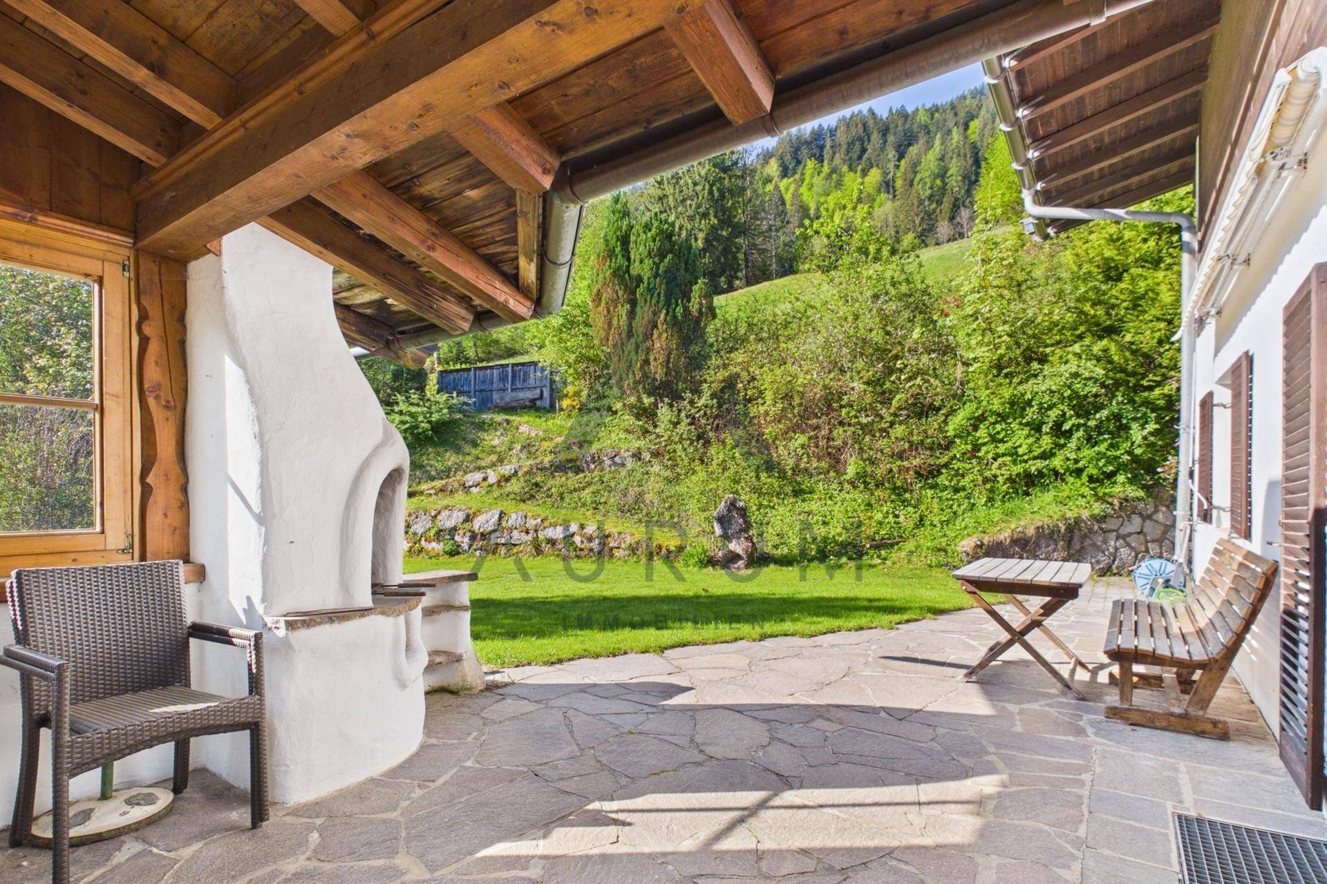Gemütliche Steinterrasse mit Außenkamin und Holzmöbeln, umgeben von einem grünen Garten und Hügeln.