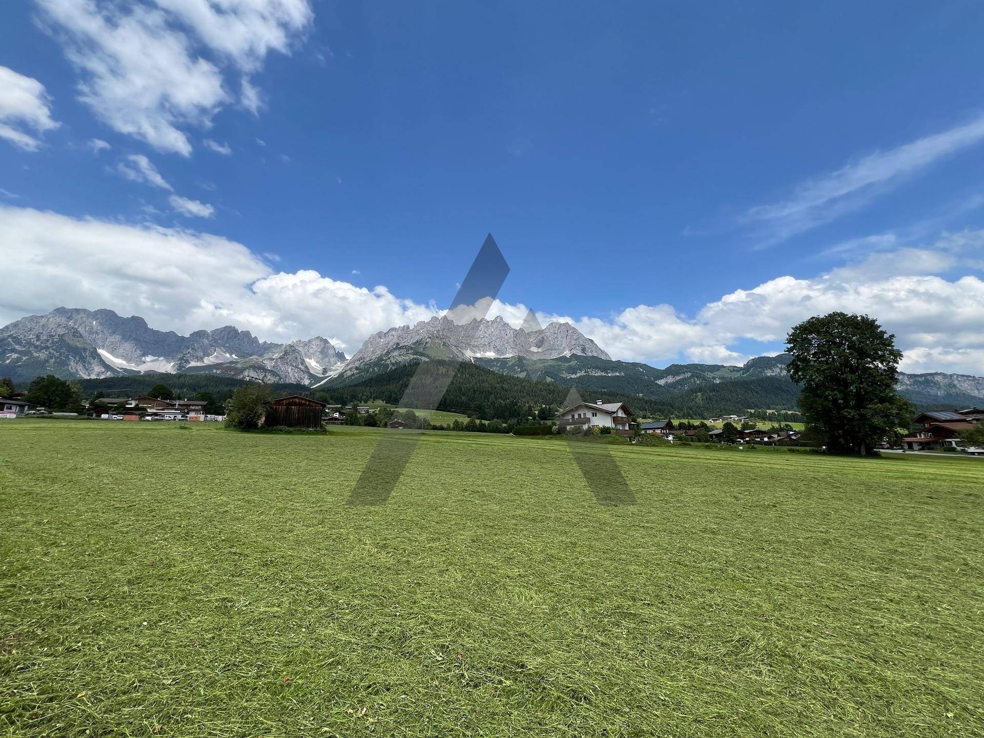 Panoramablick über grüne Felder mit einem Haus und einer majestätischen Bergkette im Hintergrund.