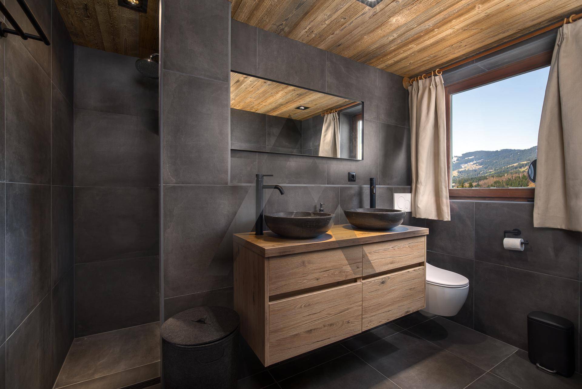 Elegantes Badezimmer mit dunklen Fliesen, Doppelwaschtisch und Bergblick durch das Fenster.