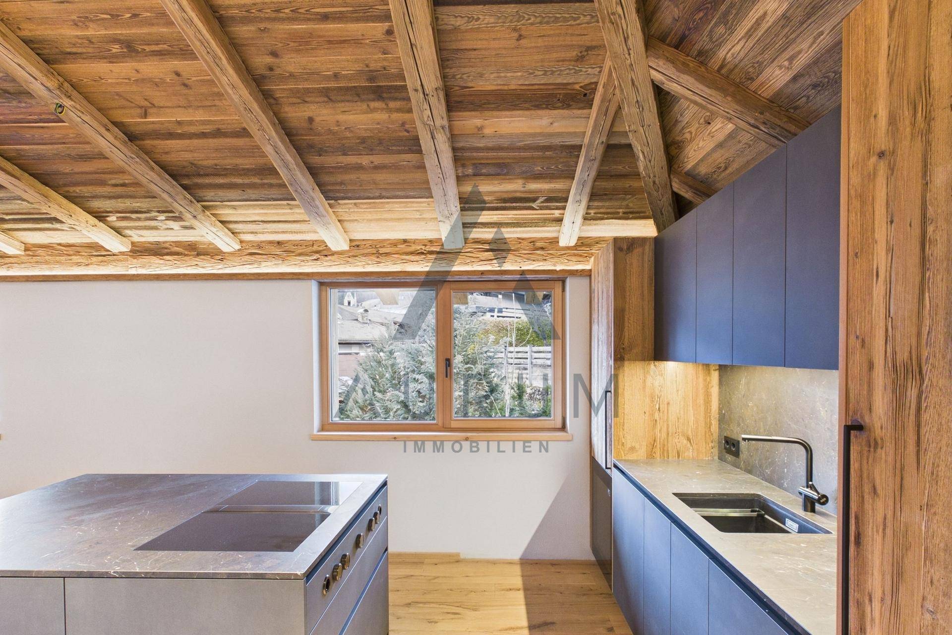 Helle Küche mit Holzbalkendecke, Kücheninsel, Induktionskochfeld und Fensterblick ins Grüne.