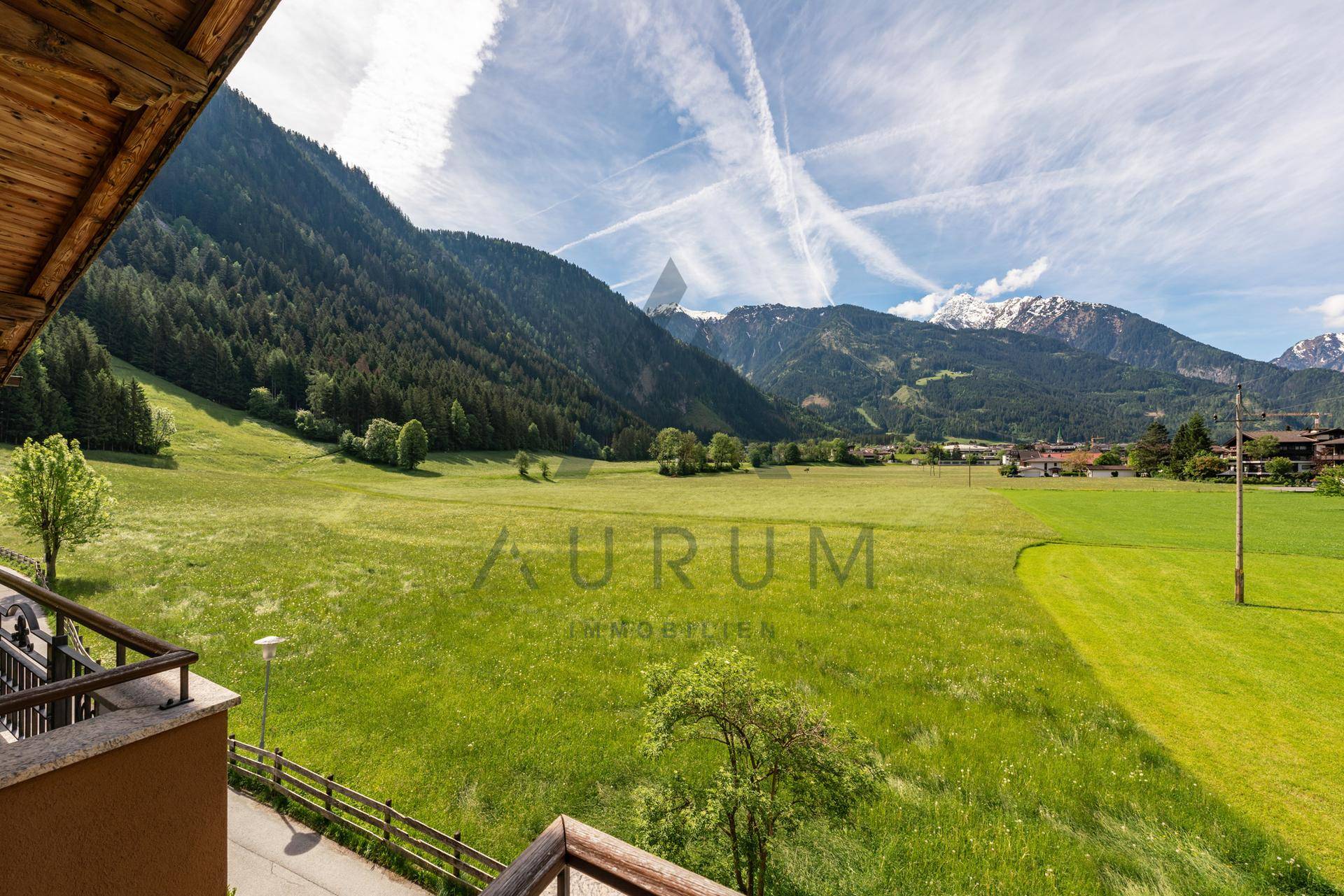 Blick von einem Balkon auf weite grüne Felder und majestätische Berge mit schneebedeckten Gipfeln unter blauem Himmel.
