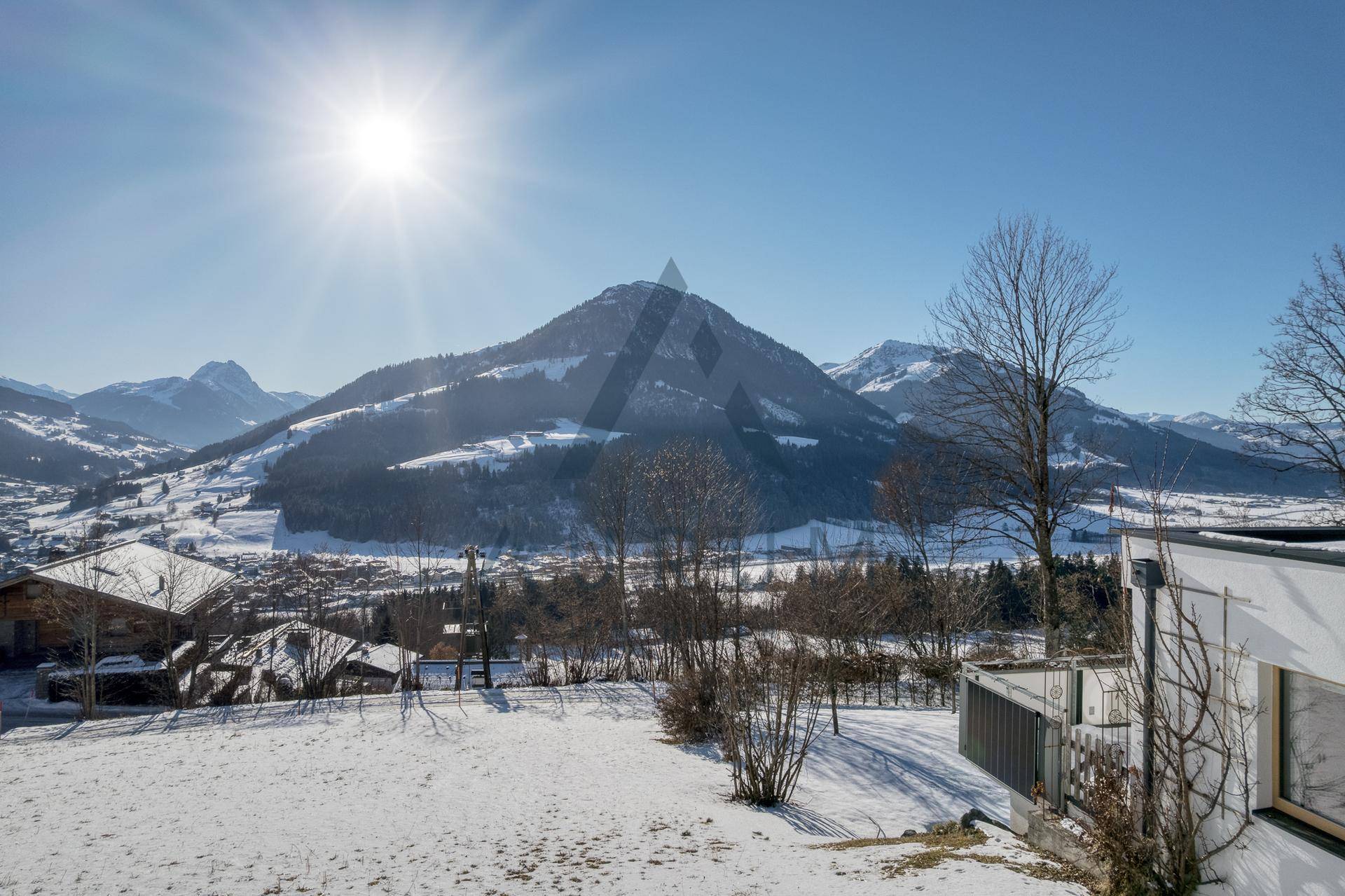 Sonniger Ausblick auf eine verschneite Landschaft mit Häusern, Bäumen und majestätischen Bergen im Hintergrund.