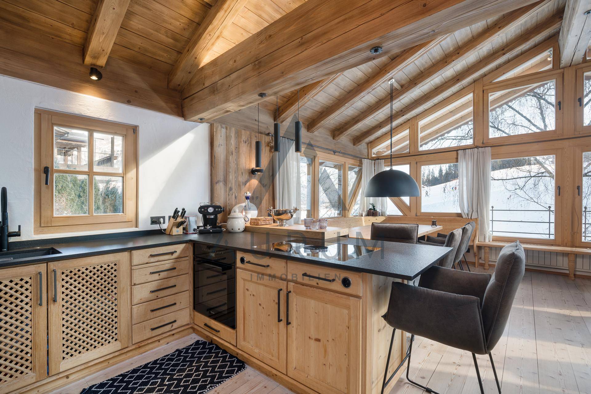Gut ausgestattete Holzküche mit Kücheninsel, modernen Geräten und viel Tageslicht durch große Fenster.