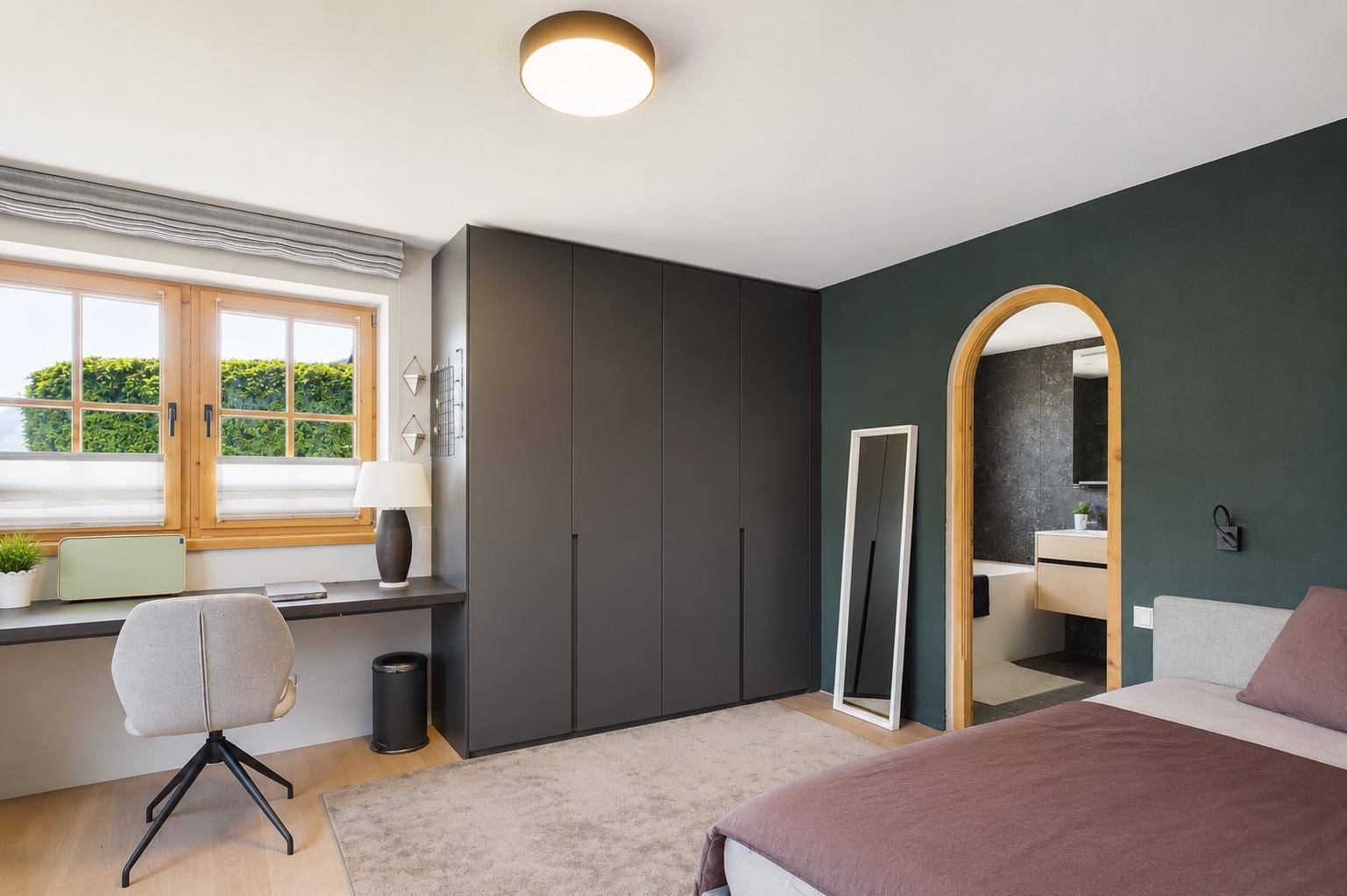 Helles Schlafzimmer mit Arbeitsbereich, Einbauschränken und Blick ins Grüne durch Holzfenster.