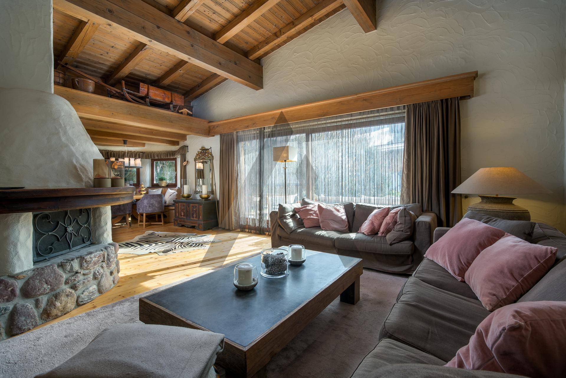 Lichtdurchflutetes Wohnzimmer mit Kamin, Holzbalken und großem Fenster mit Blick nach draußen.