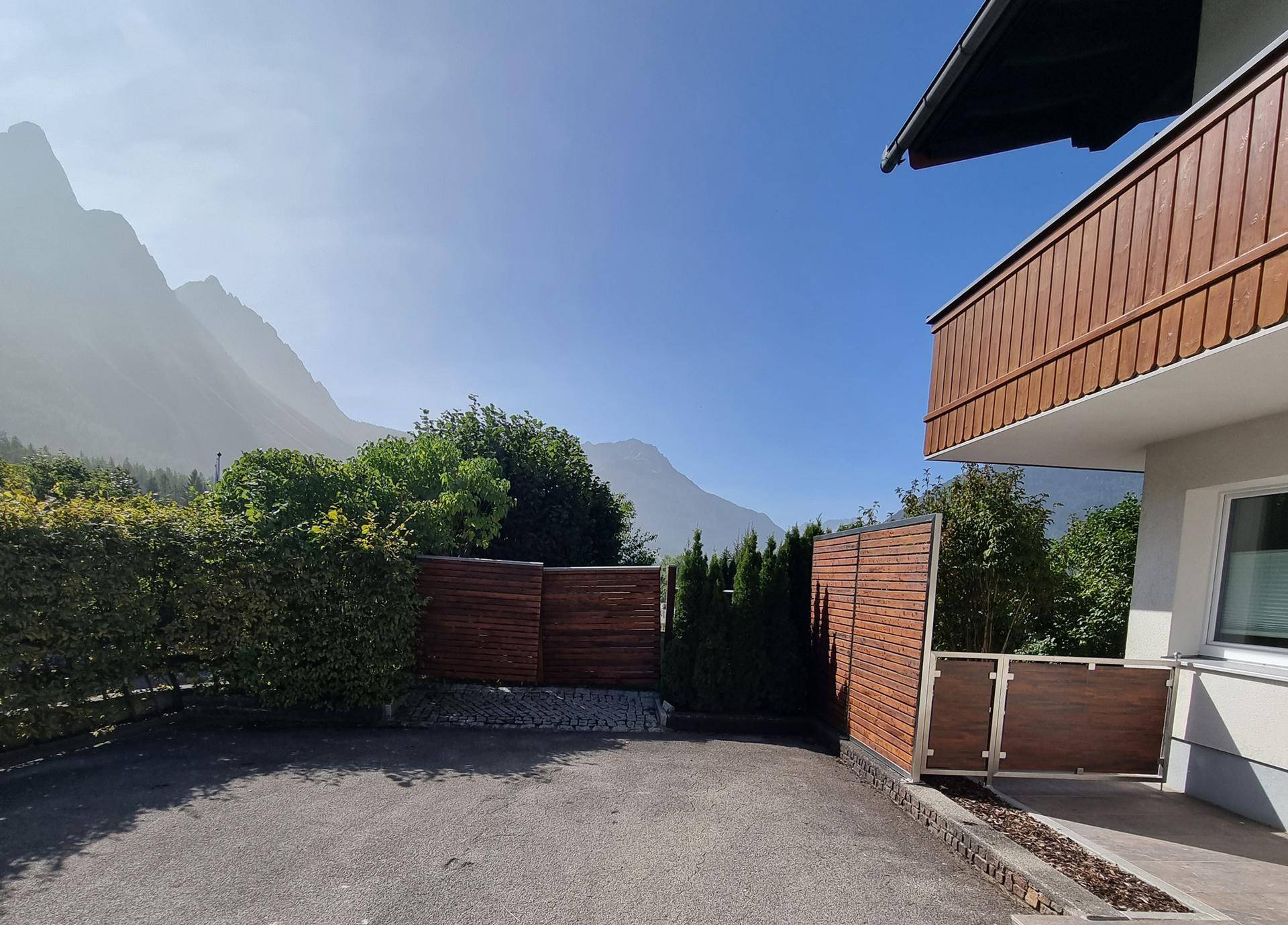 Seitenansicht des Hauses mit Holzbalkon, Hecke und beeindruckender Bergkulisse.