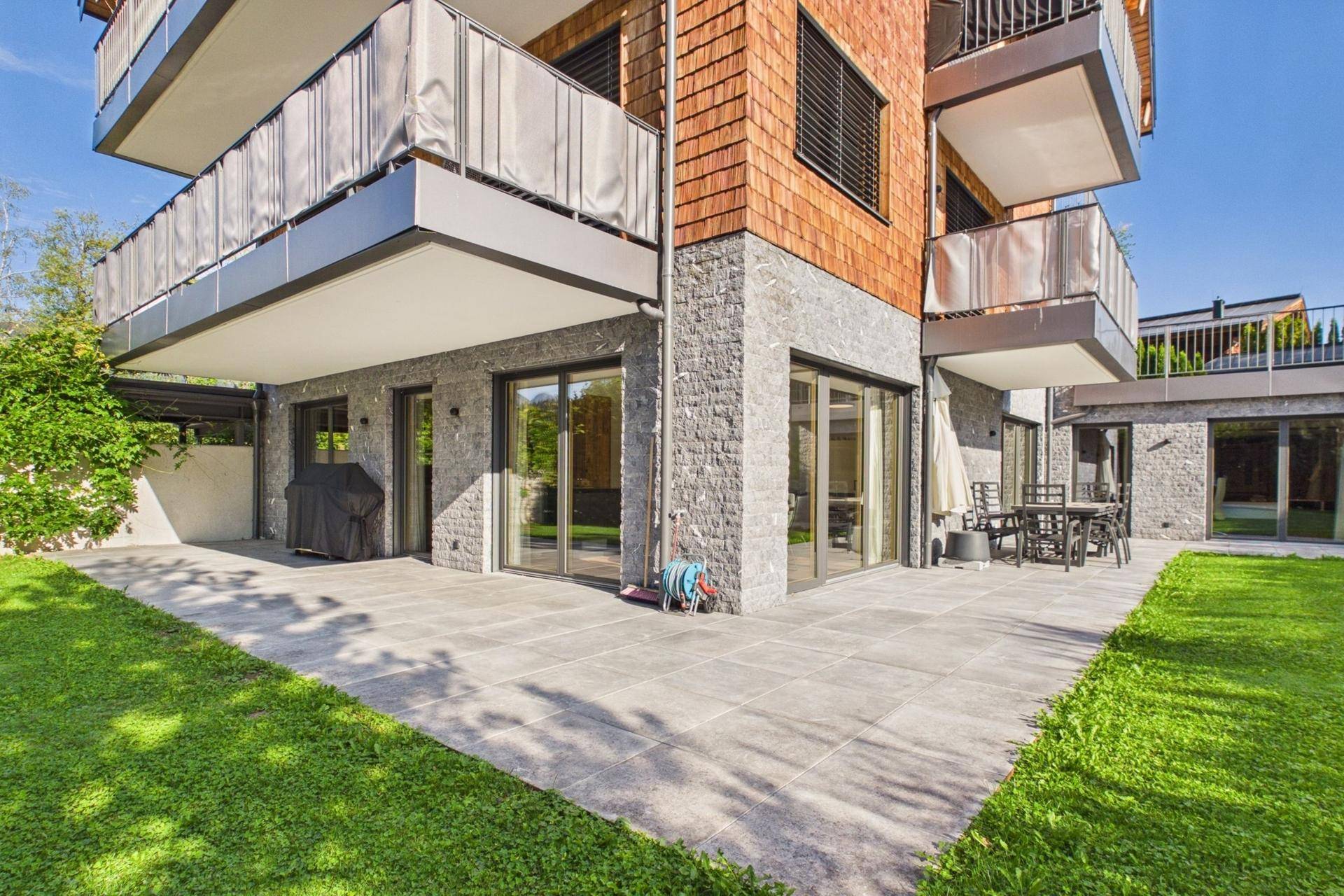 Außenbereich mit großer gepflasterter Terrasse, Rasenfläche und Balkonen am modernen Gebäude.