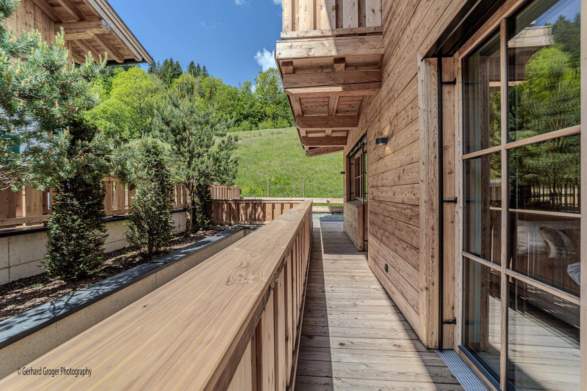 Langer Holzbalkon mit Geländer und Zugang zu den Zimmern, umgeben von grüner Natur.