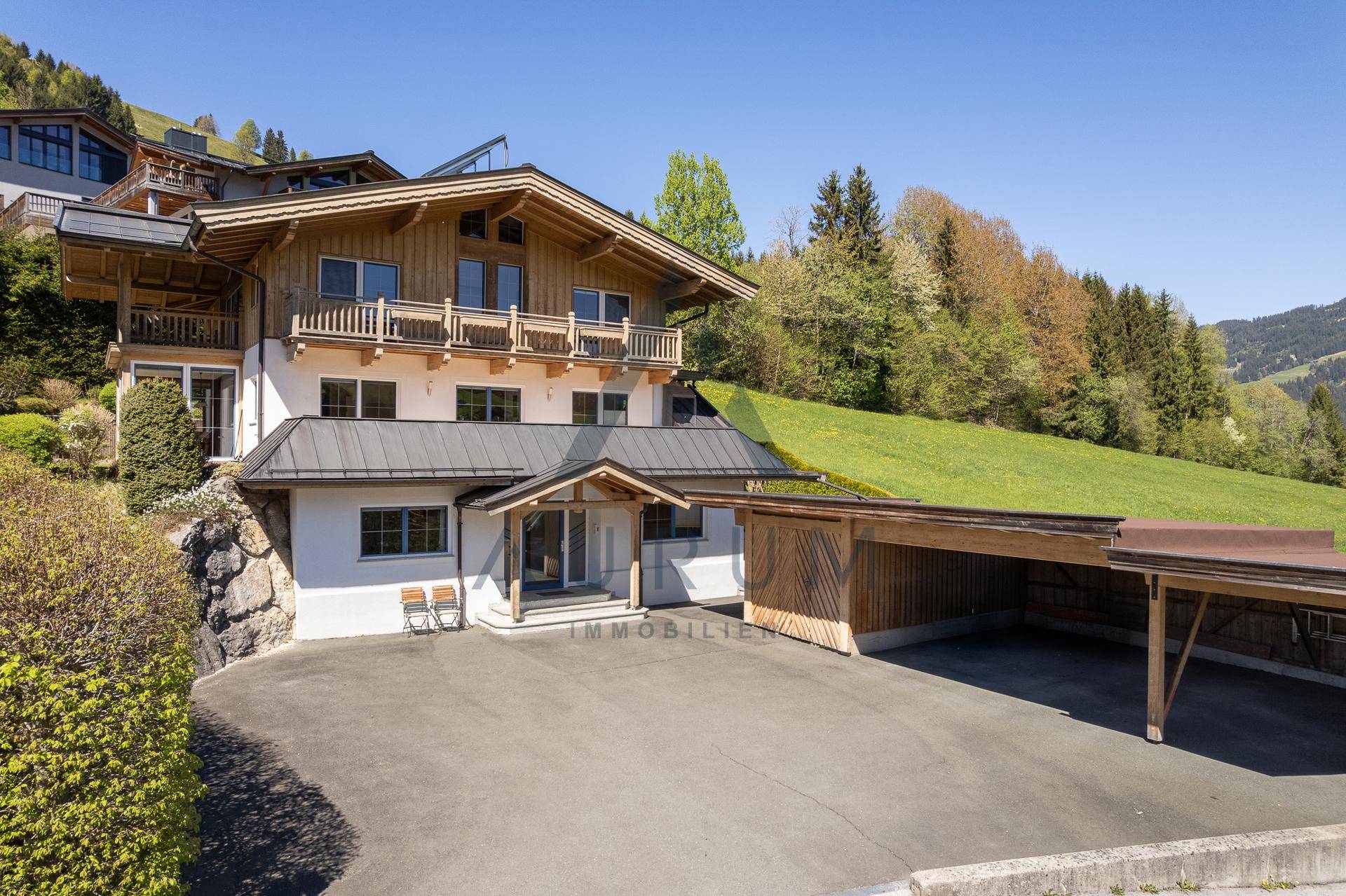 Traditionelles Holzhaus mit Balkonen und einem Carport, eingebettet in eine grüne Berglandschaft.