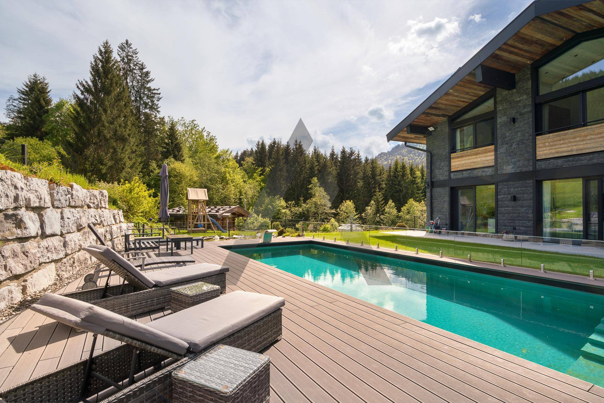 Entspannender Poolbereich mit Holzdeck und Liegestühlen, umgeben von Wald und Bergen.