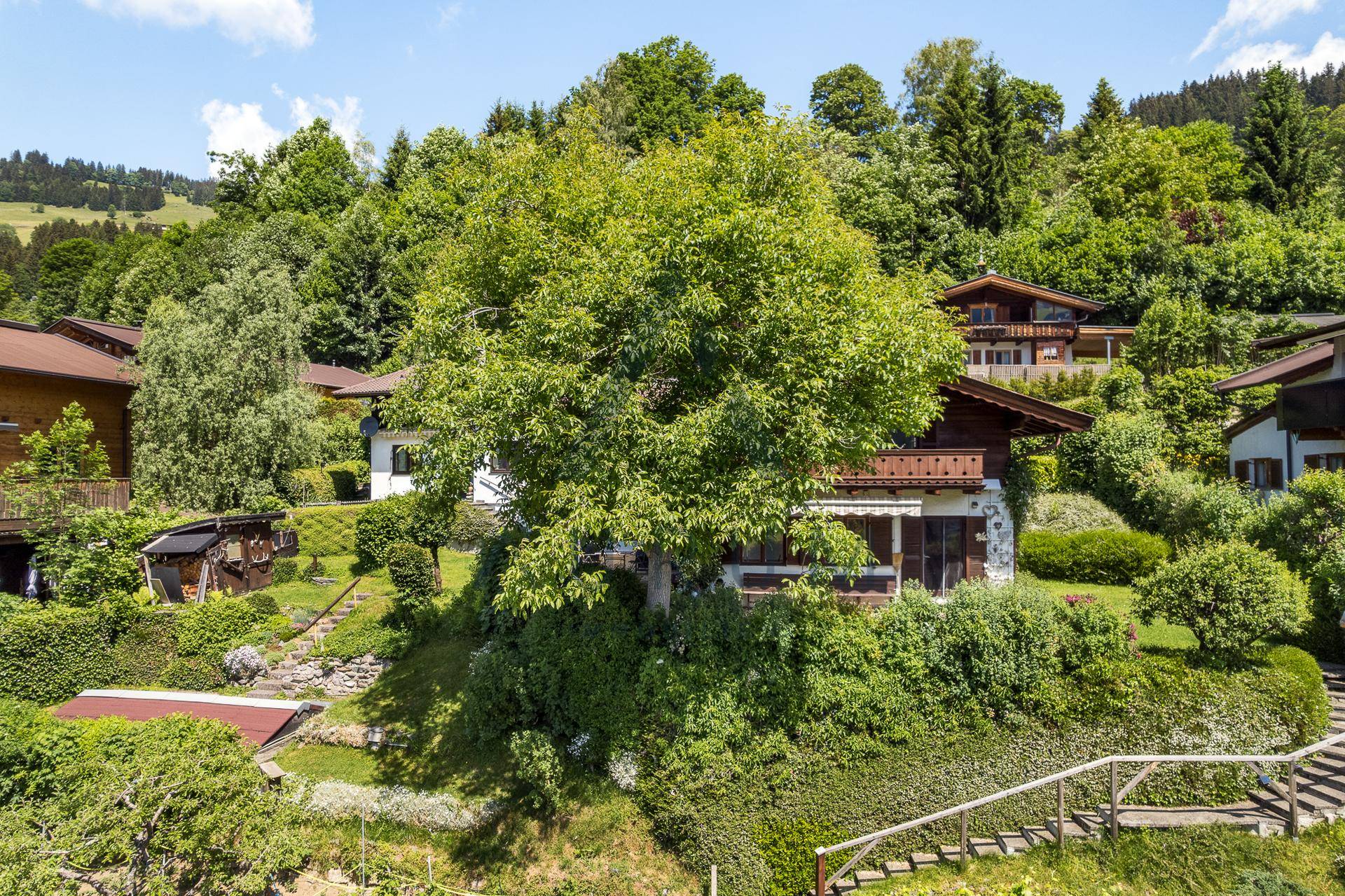 Idyllisches Haus mit üppigem Garten und altem Baumbestand, umgeben von grünen Hügeln.