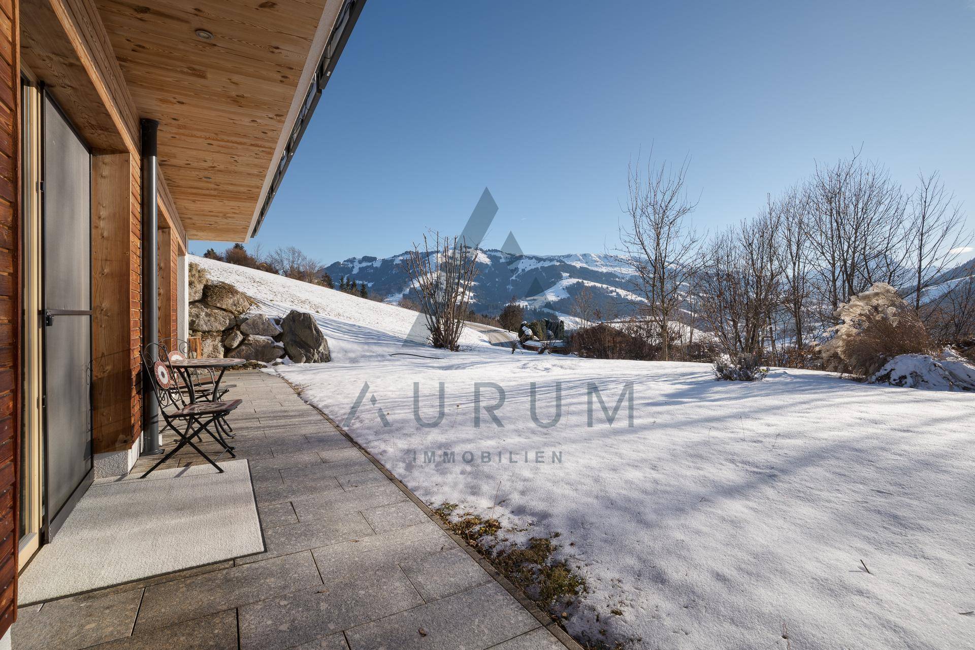 Gepflasterte Terrasse neben dem Haus, umgeben von Schnee und einem weitläufigen Garten mit Bergblick.